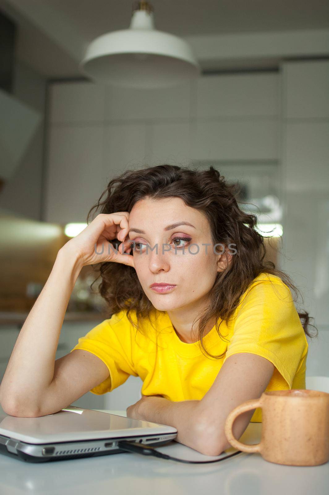 Tired young woman with curly hair and yellow shirt is working from home using her laptop at the kitchen table in her apartment, remote work, freelance, burnout syndrome by balinska_lv