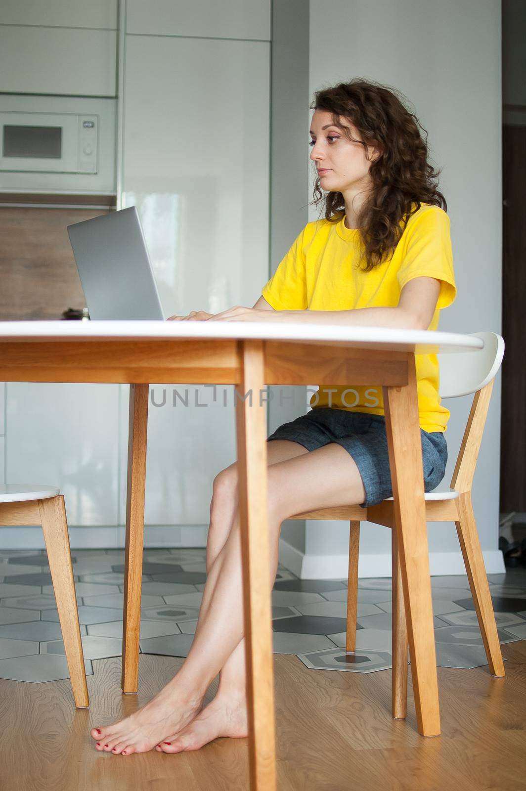 Young woman with curly hair and yellow shirt is working from home using her laptop at the kitchen table in her apartment, remote work, freelance by balinska_lv