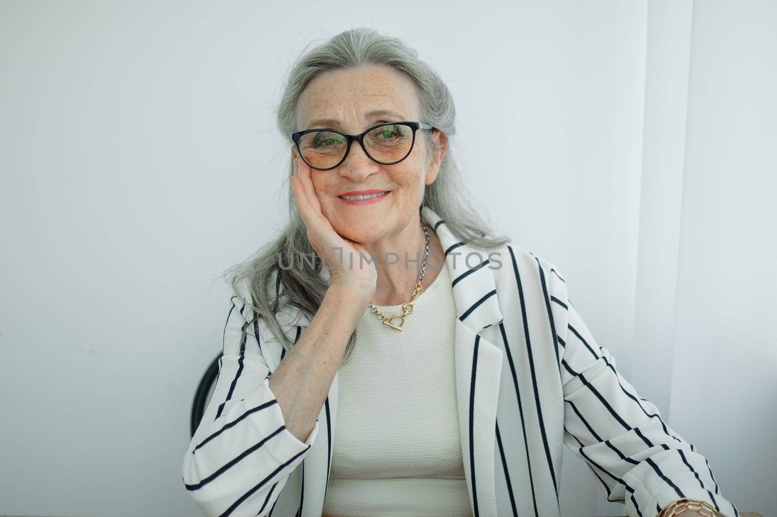 Senior grey haired businesswoman in striped jacket with eyeglasses is working in her office sitting at the desk. Happy retirement, employment and labor by balinska_lv