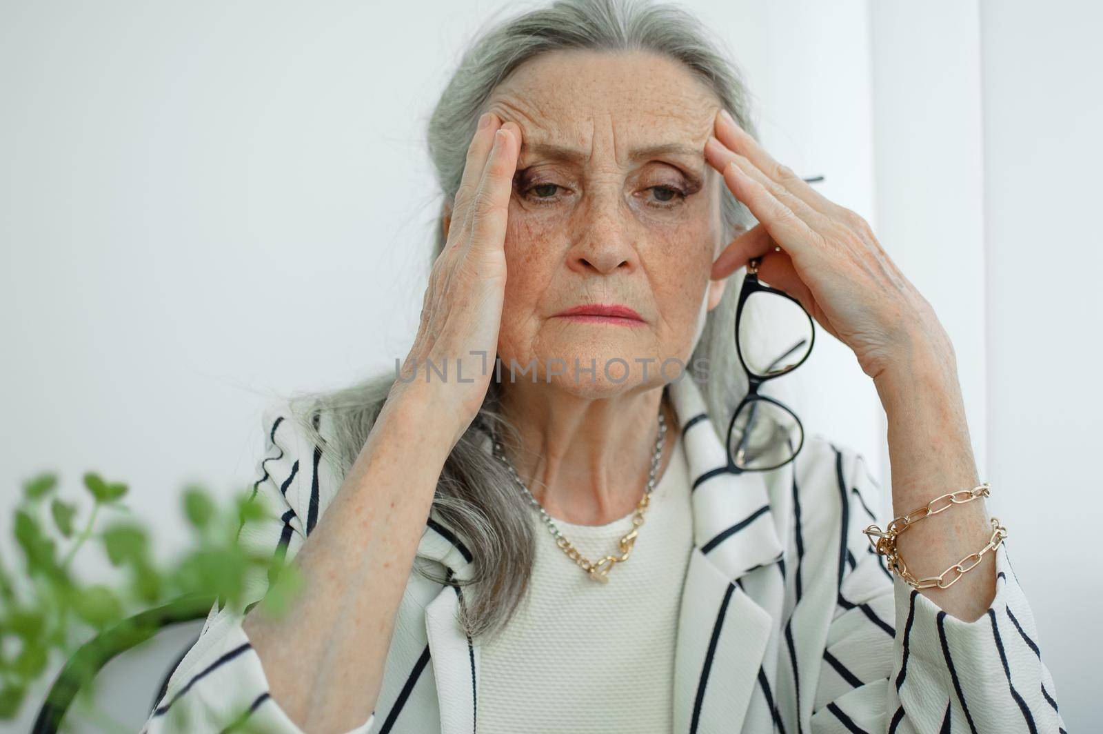 Tired senior grey haired businesswoman in striped jacket with eyeglasses is working in her office sitting at the desk and feeling bad due to menopause, menopause relief concept by balinska_lv