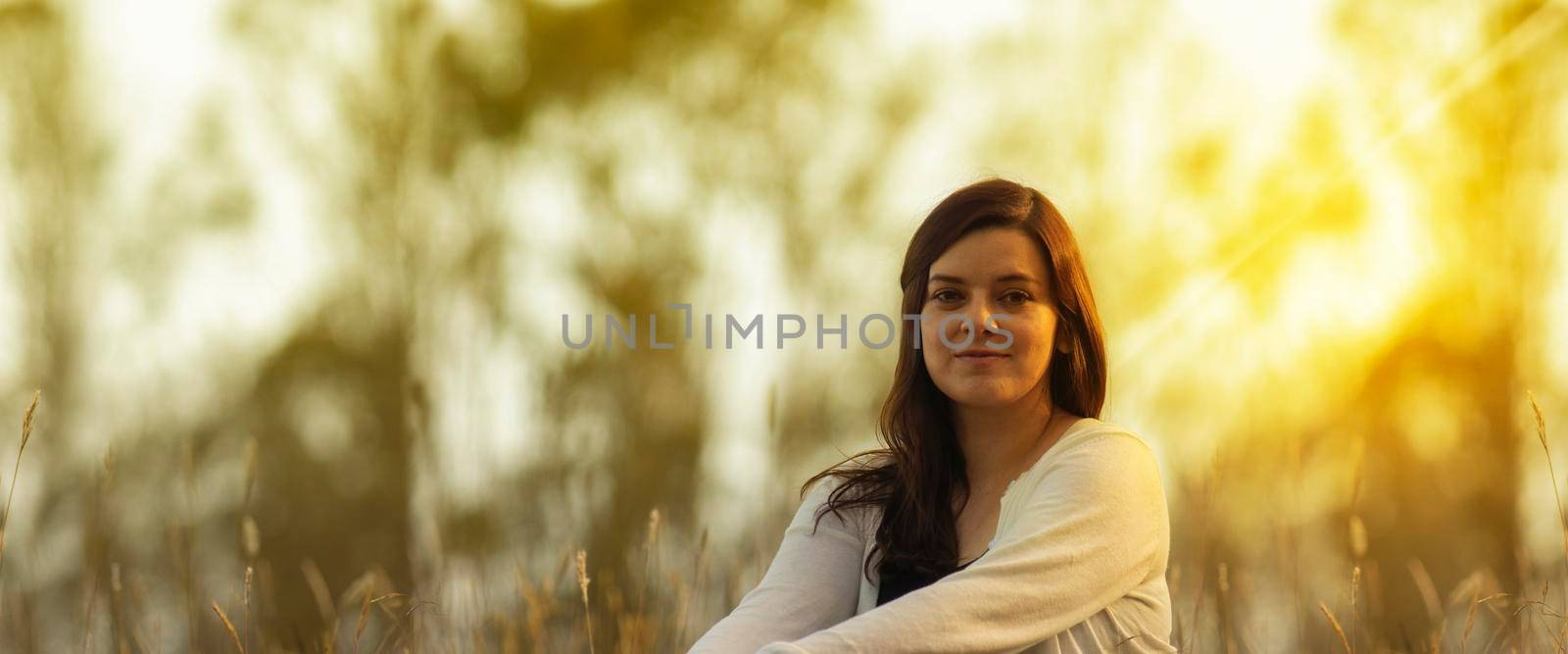 Portrait of beautiful Hispanic young woman with long hair looking at the camera against a background of unfocused trees during sunset by alejomiranda