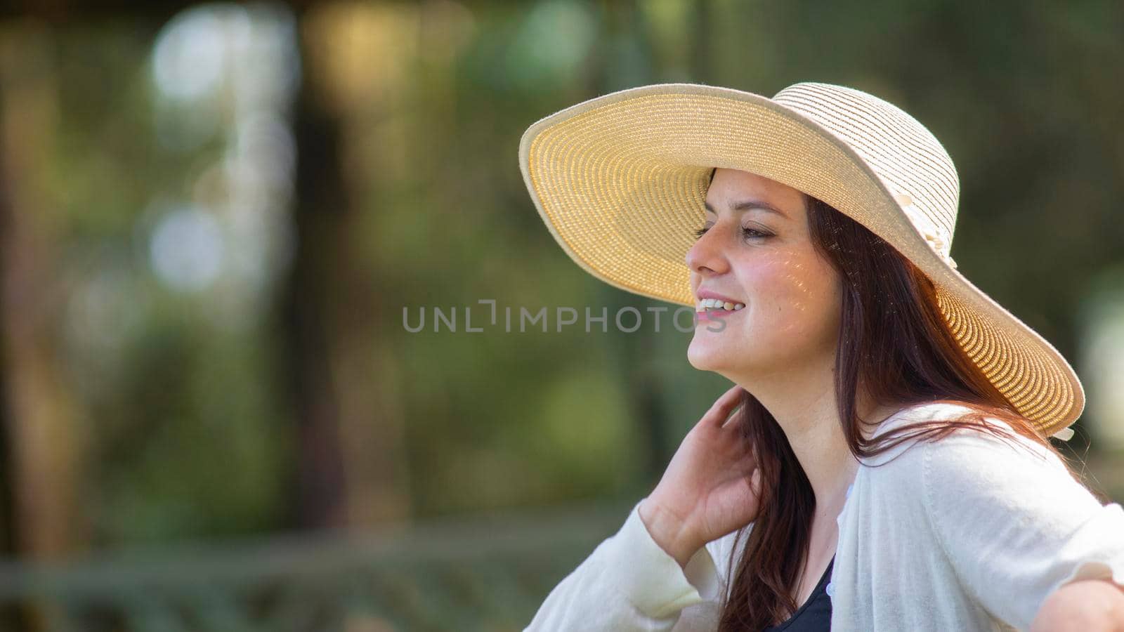 Portrait of beautiful young Hispanic woman with long hair with a hat very cheerful sitting on a park bench against a background of unfocused green trees by alejomiranda