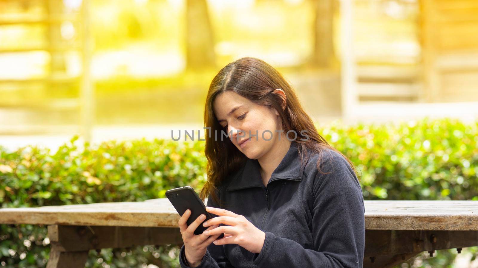 Beautiful Hispanic young woman reading a message on her cell phone sitting on a park bench during the morning