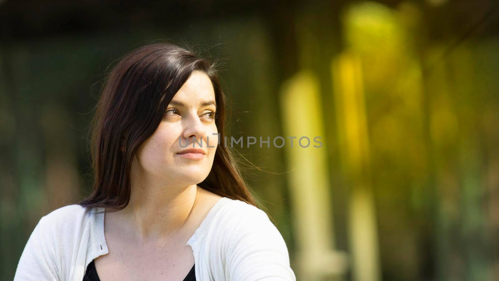 Portrait of beautiful Hispanic young woman with long hair looking to the right against a background of unfocused trees during sunset with copy space