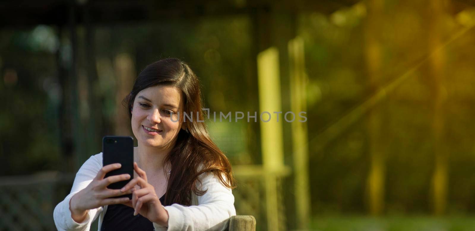 Beautiful Hispanic young woman reading a message on her cell phone sitting on a park bench  against a background of unfocused green trees during sunset