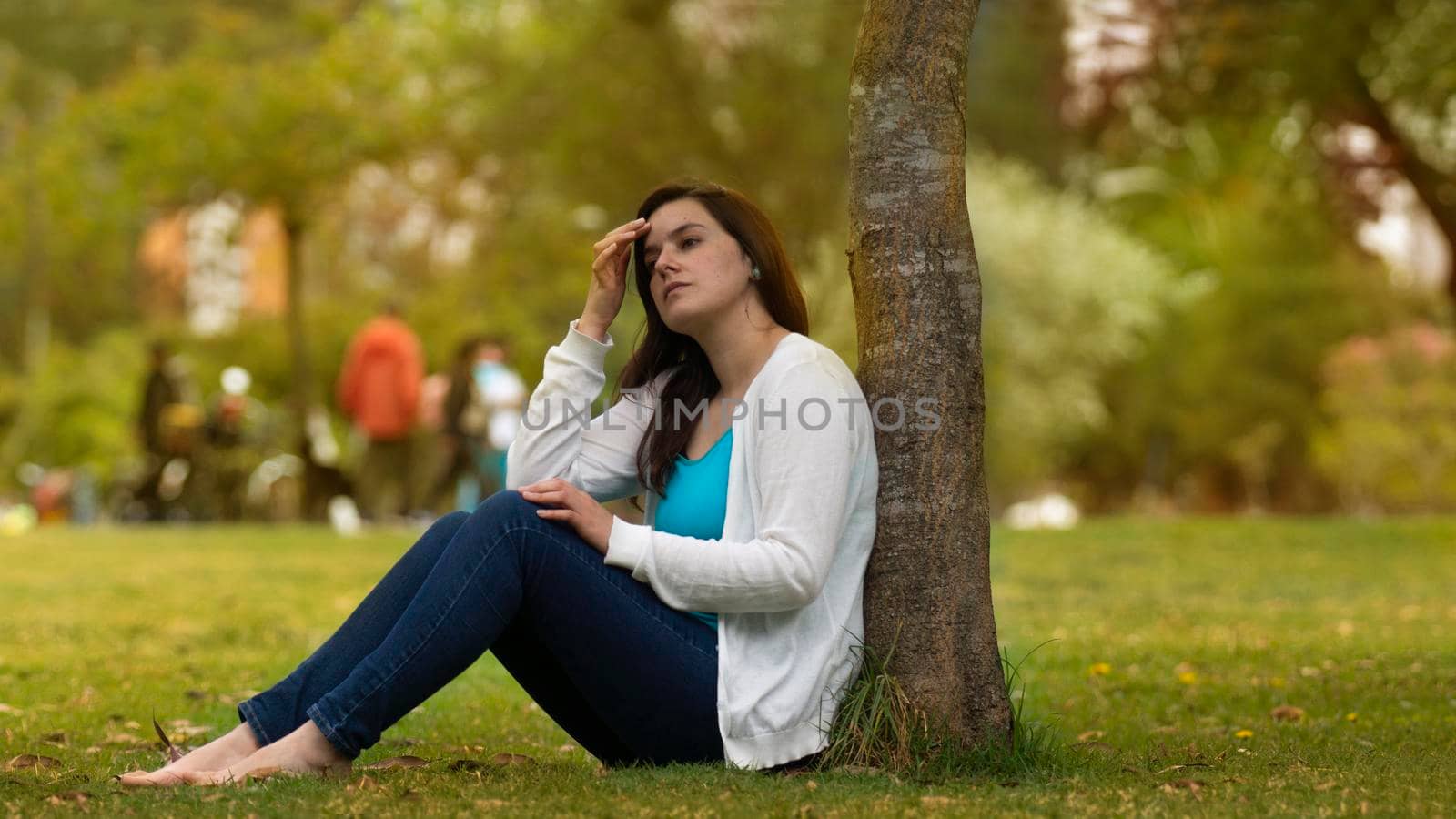 Portrait of beautiful young Hispanic woman sitting under a tree in the middle of a park with a pensive attitude against a background of unfocused trees during the day