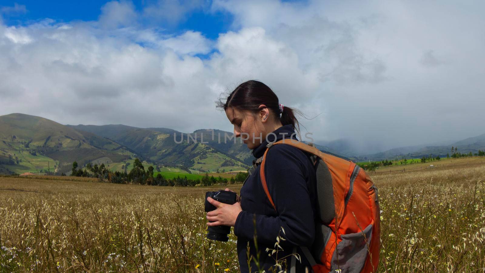 Beautiful Hispanic female explorer seen from behind with backpack reviewing photos on her camera in the middle of a sown field on a cloudy morning