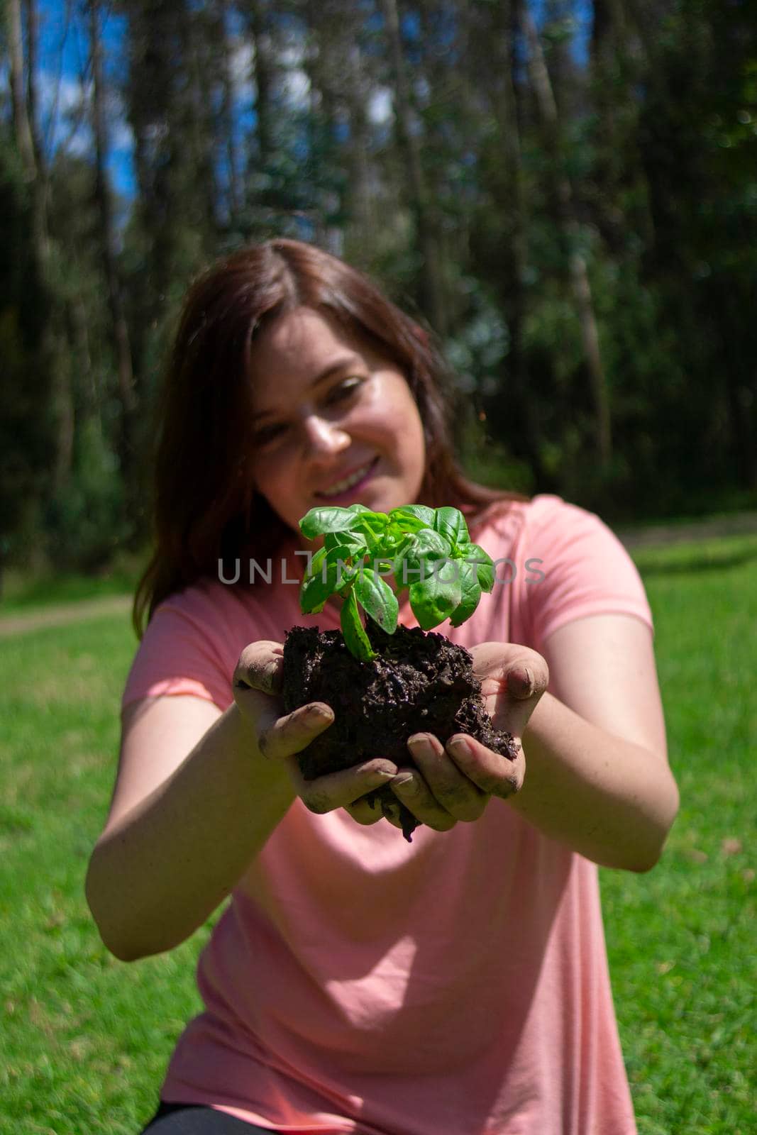 Beautiful young Hispanic woman holding a small plant in her field hands before being planted in a green field surrounded by trees during the morning