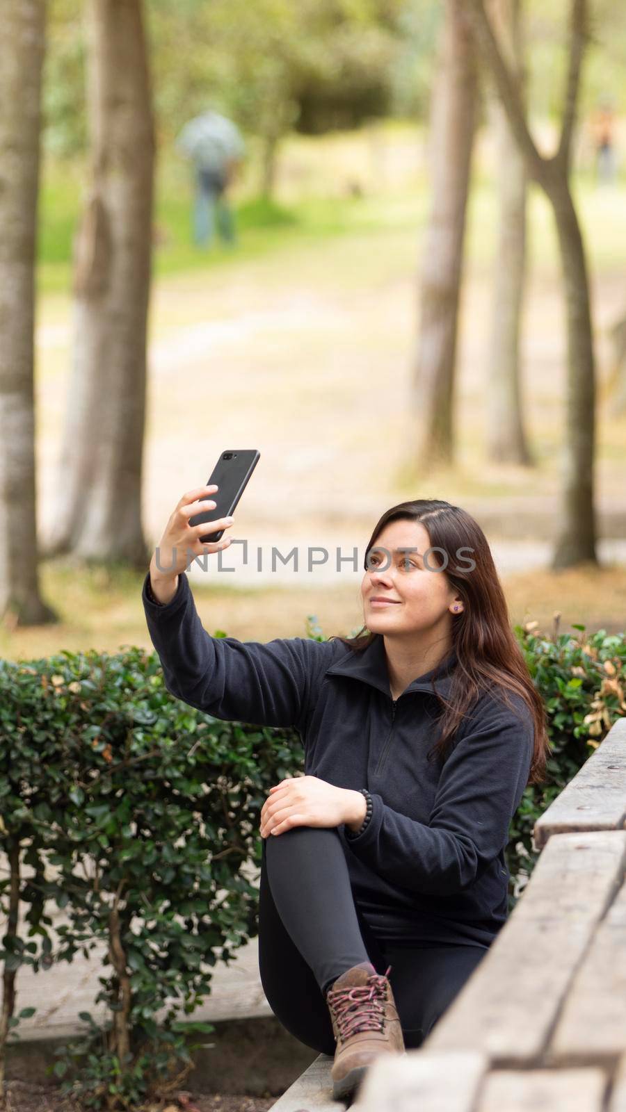 Beautiful young Hispanic woman taking a selfie with her cell phone sitting on a park bench by alejomiranda