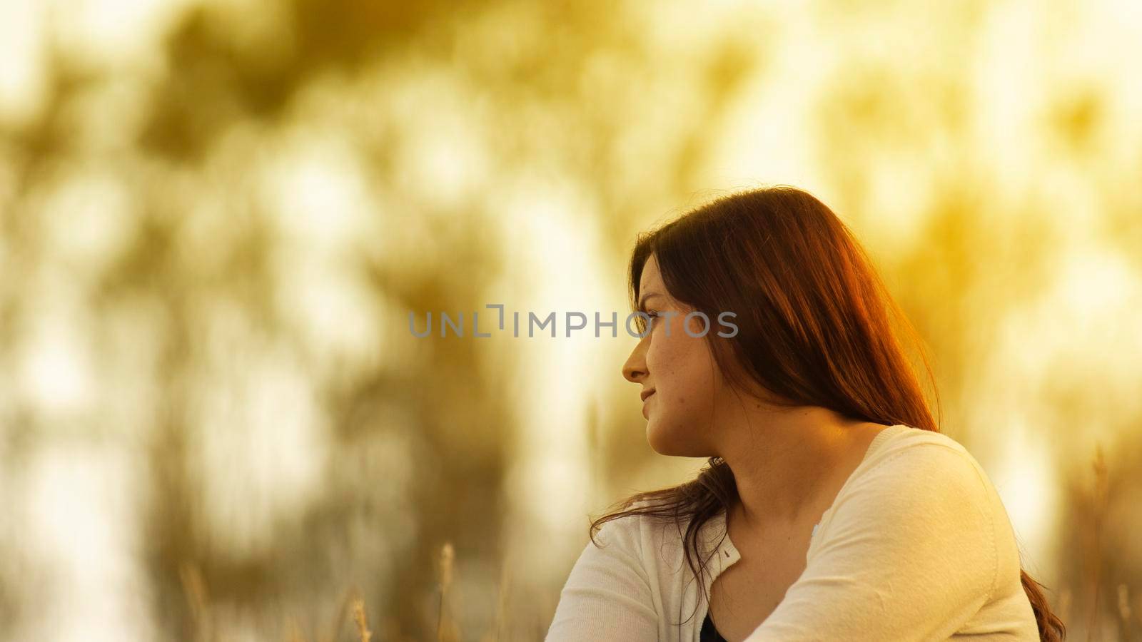 Portrait of beautiful Hispanic young woman with long hair looking to the right against a background of unfocused trees during sunset with copy space