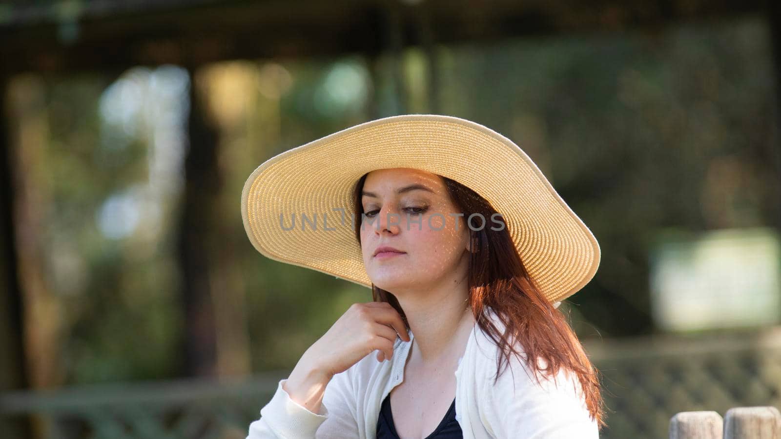 Portrait of beautiful long-haired Hispanic young woman wearing a hat sitting on a park bench with a pensive attitude against a background of unfocused green trees during sunset