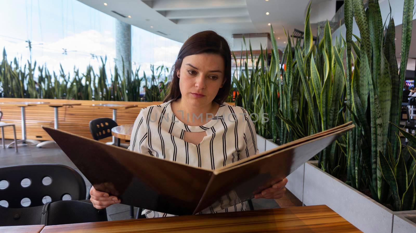 Close up on a beautiful Hispanic woman reading the menu sitting in front of a table in a restaurant adorned with plants