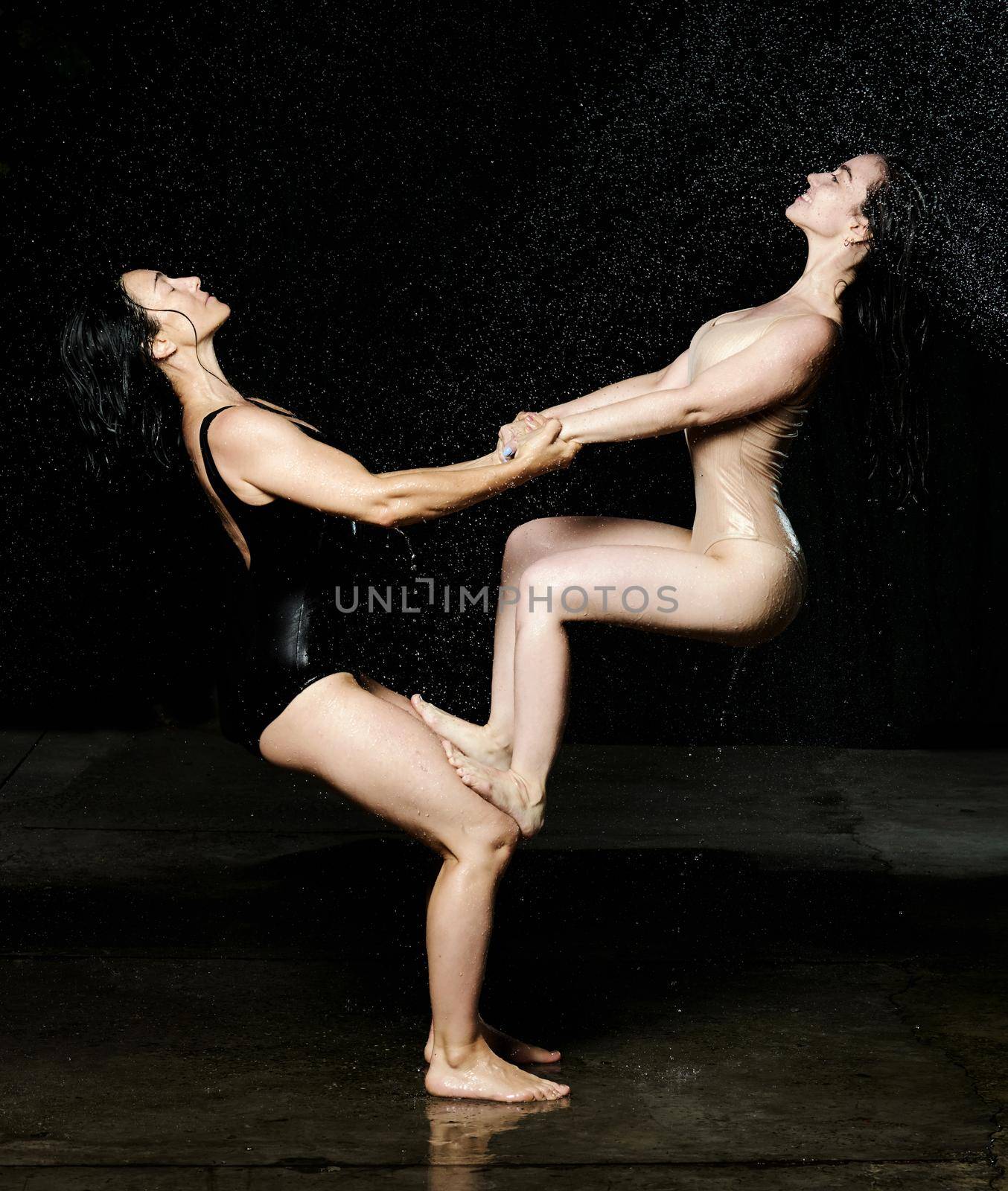 two women of Caucasian appearance perform an acrobatic stunt on a black background with rain. Woman holds on her knees another in beige bodysuit by ndanko