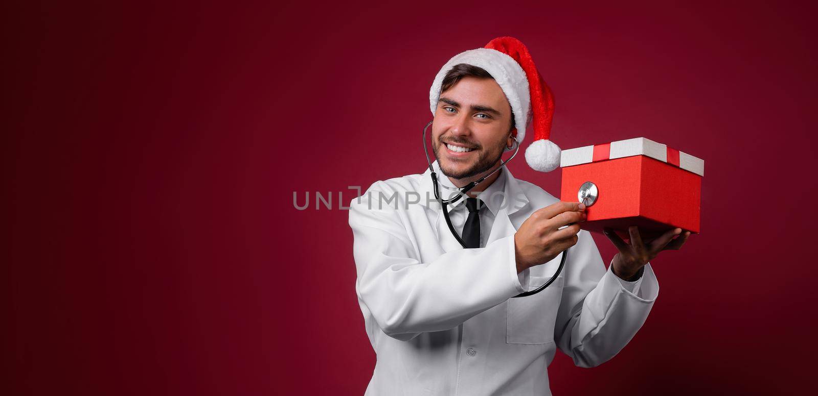 Young handsome doctor in white uniforme and Santa Claus hat standing in studio on red background smile and finger in camera by andreonegin
