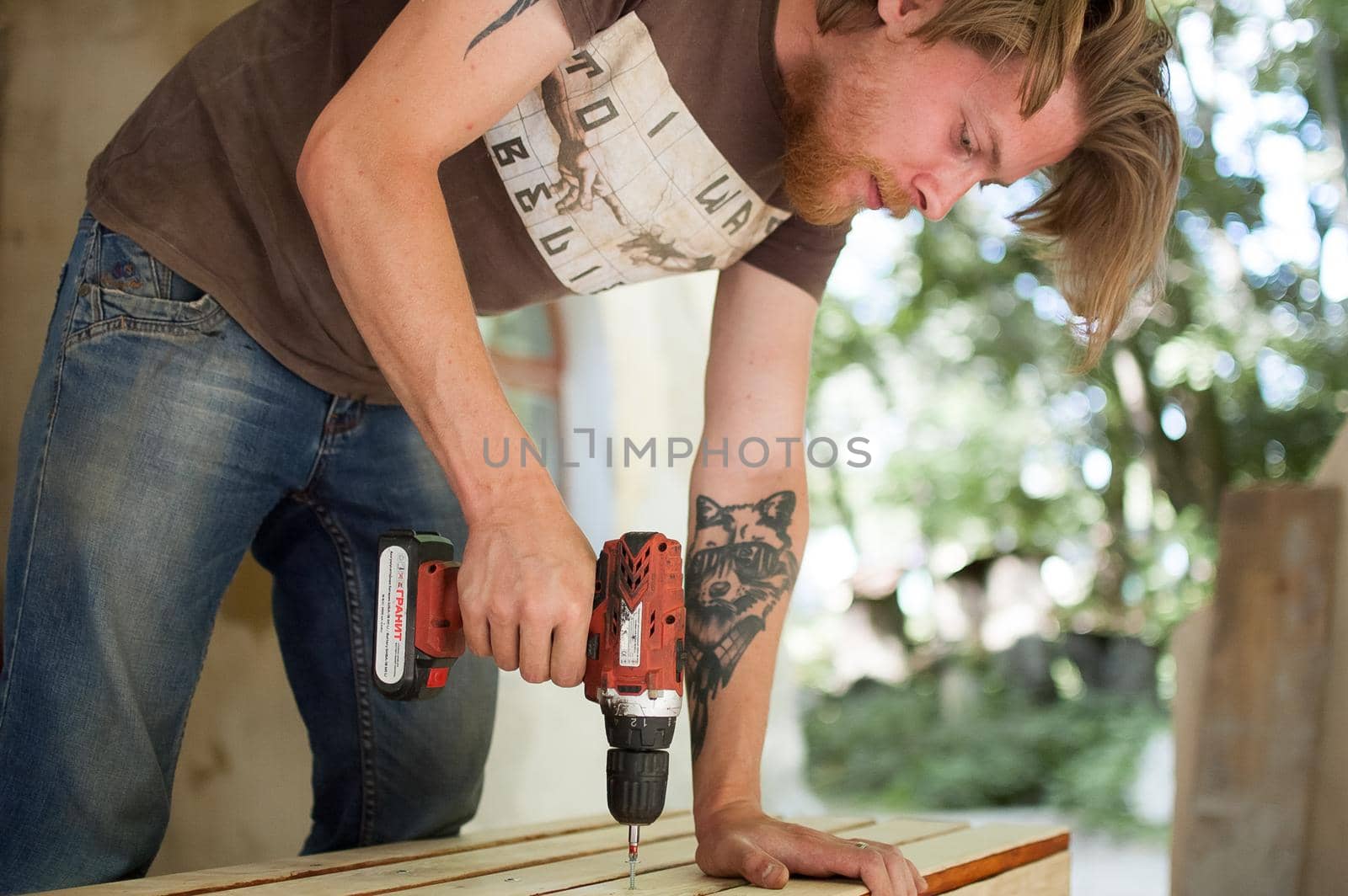 13.08.2021 - Ukraine, Goshcha, voluntary event, skilled young male worker is using power screwdriver drilling during construction wooden bench, do it yourself.