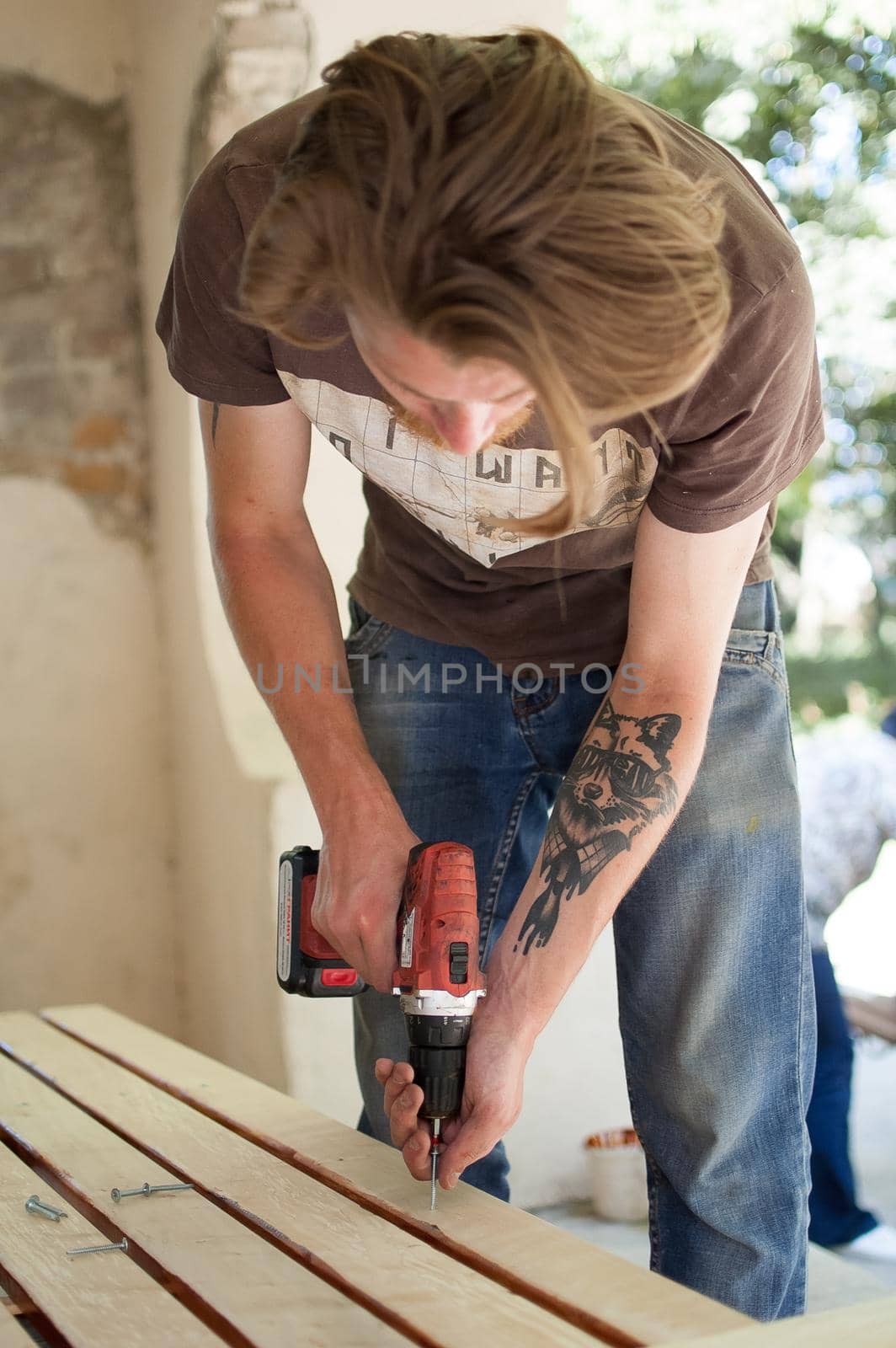 13.08.2021 - Ukraine, Goshcha, voluntary event, skilled young male worker is using power screwdriver drilling during construction wooden bench, do it yourself.