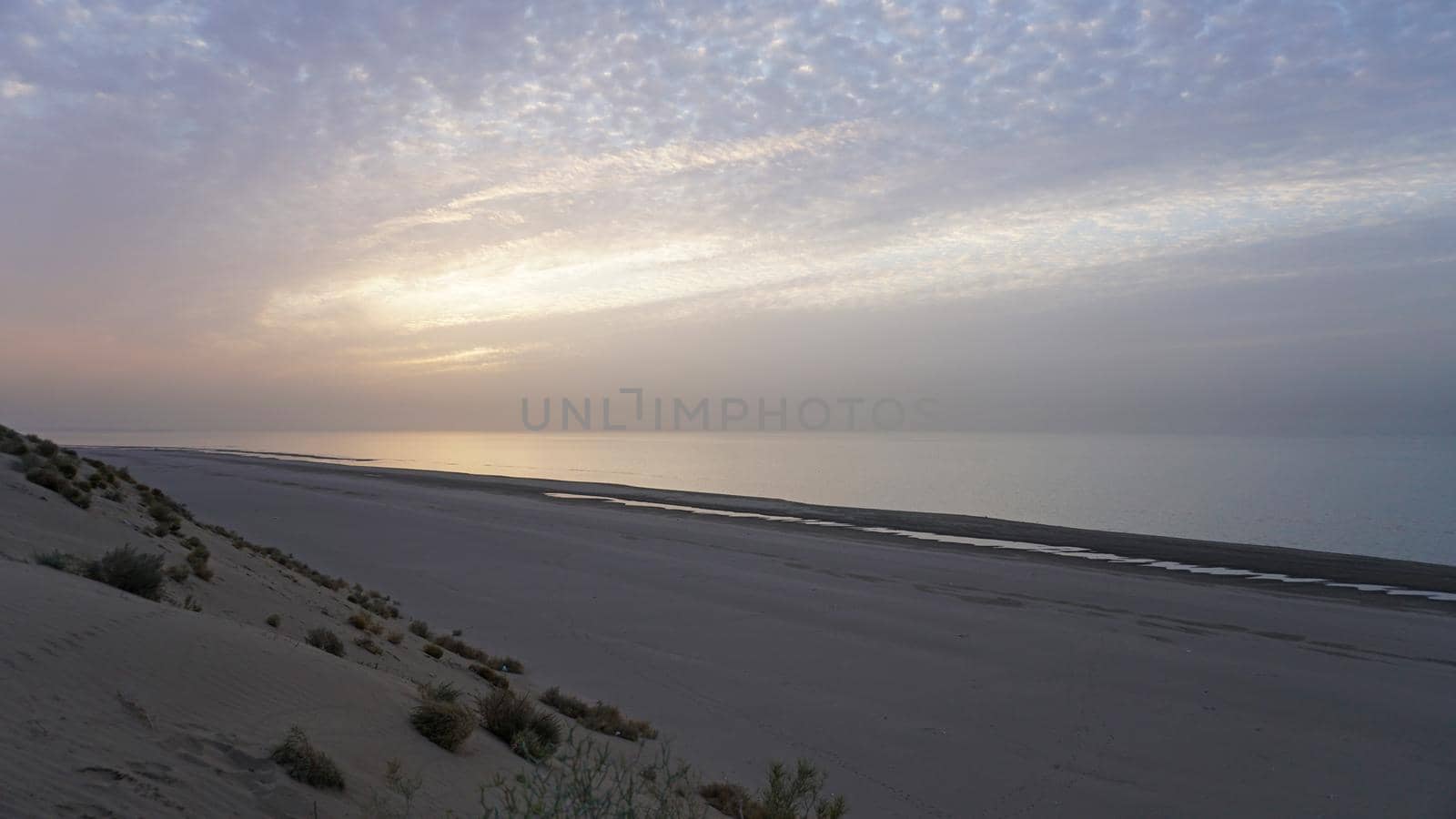 Dawn on the sandy beach of the sea. Sand dune. The sun's rays peek out from behind the clouds. Car tracks in the sand. A light wave, almost calm. The water merges with the sky. Autumn morning