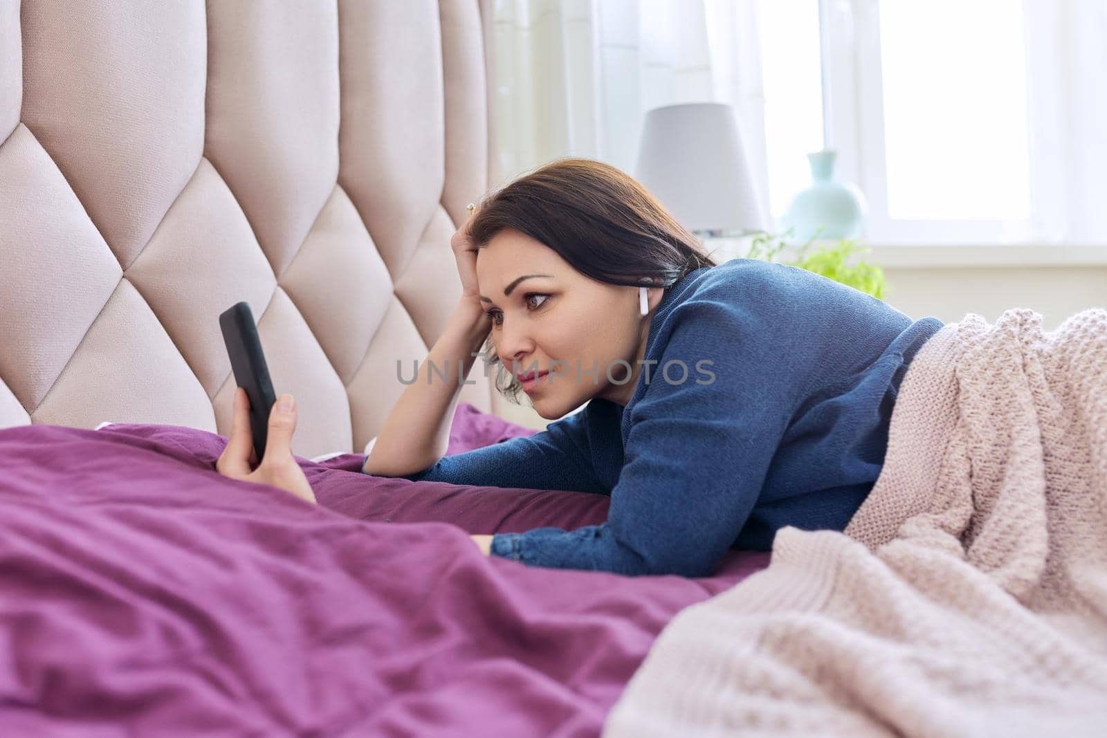 Smiling middle-aged woman in headphones looking in smartphone, resting female lying in bed at home, video on the phone, film, vlog, social network. Lifestyle, rest, technology, mature people concept