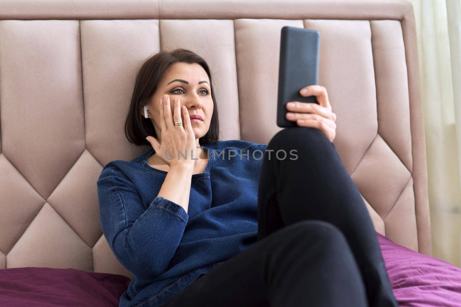 Sad middle aged woman in headphones looking into the smartphone screen. A frustrated female sitting at home in bed watching an unpleasant video. Technology, emotions, lifestyle, mature age concept