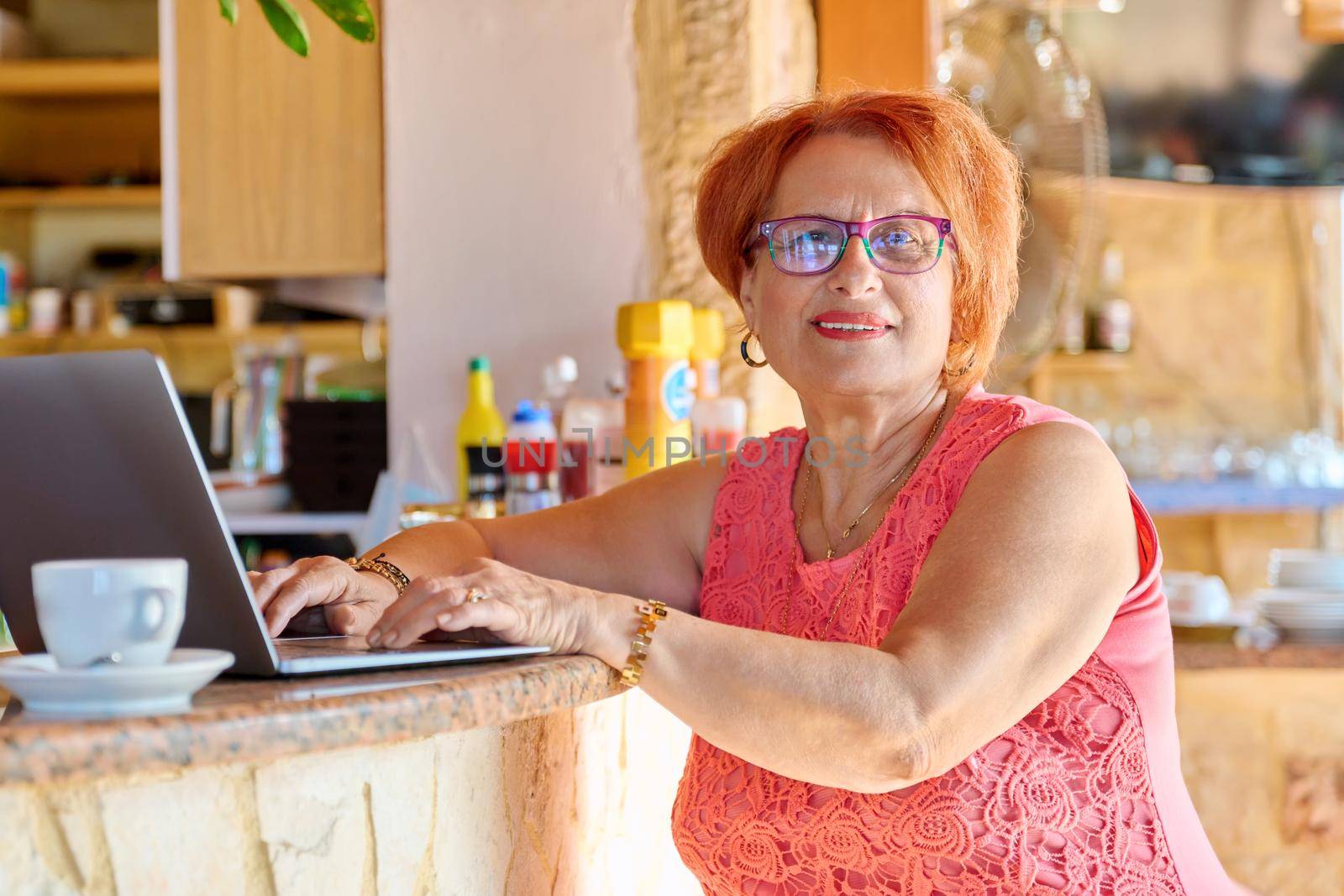 Smiling positive elderly woman looking at laptop screen. A female of retirement age in a cafe with a laptop. Active lifestyle of retirees, technology, family business, 70s people concept