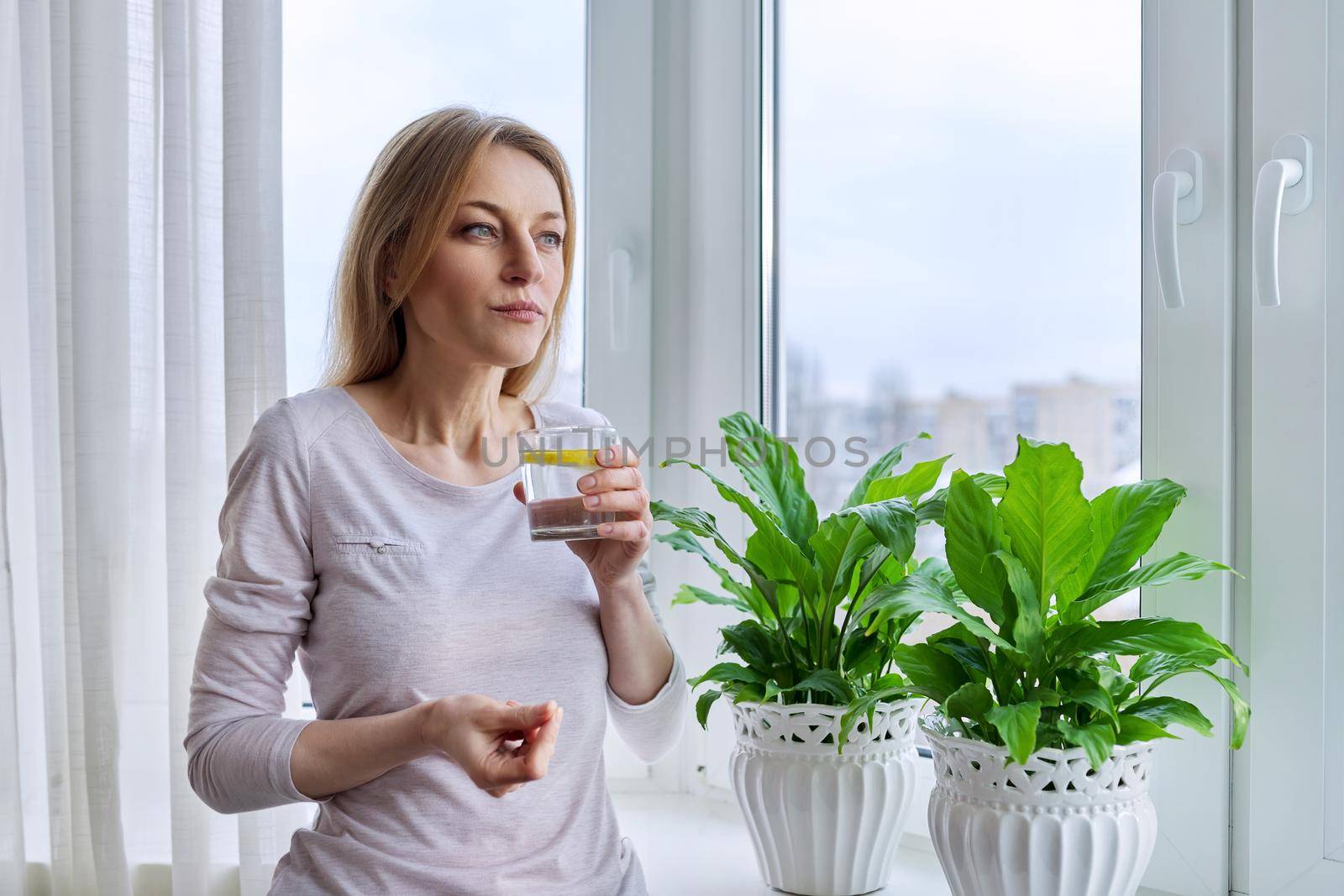 Middle-aged woman drinking lemon water at home near the window by VH-studio