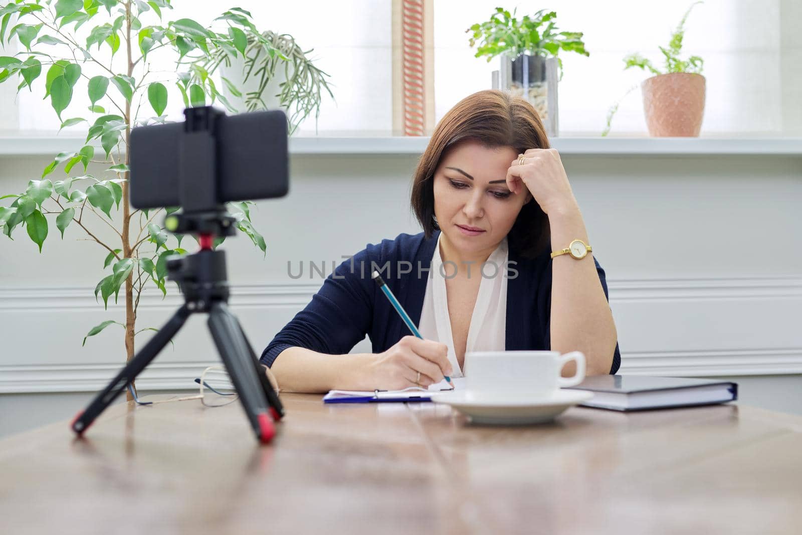 Middle-aged woman teacher, business woman working online using smartphone. Business confident female with papers on table talking looking in webcam phone on tripod, at home. Technology education work