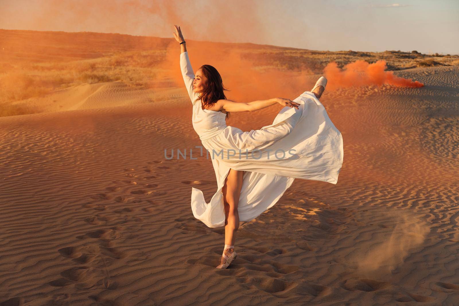 A girl in a fly white dress dances and poses in the sand desert at sunset.