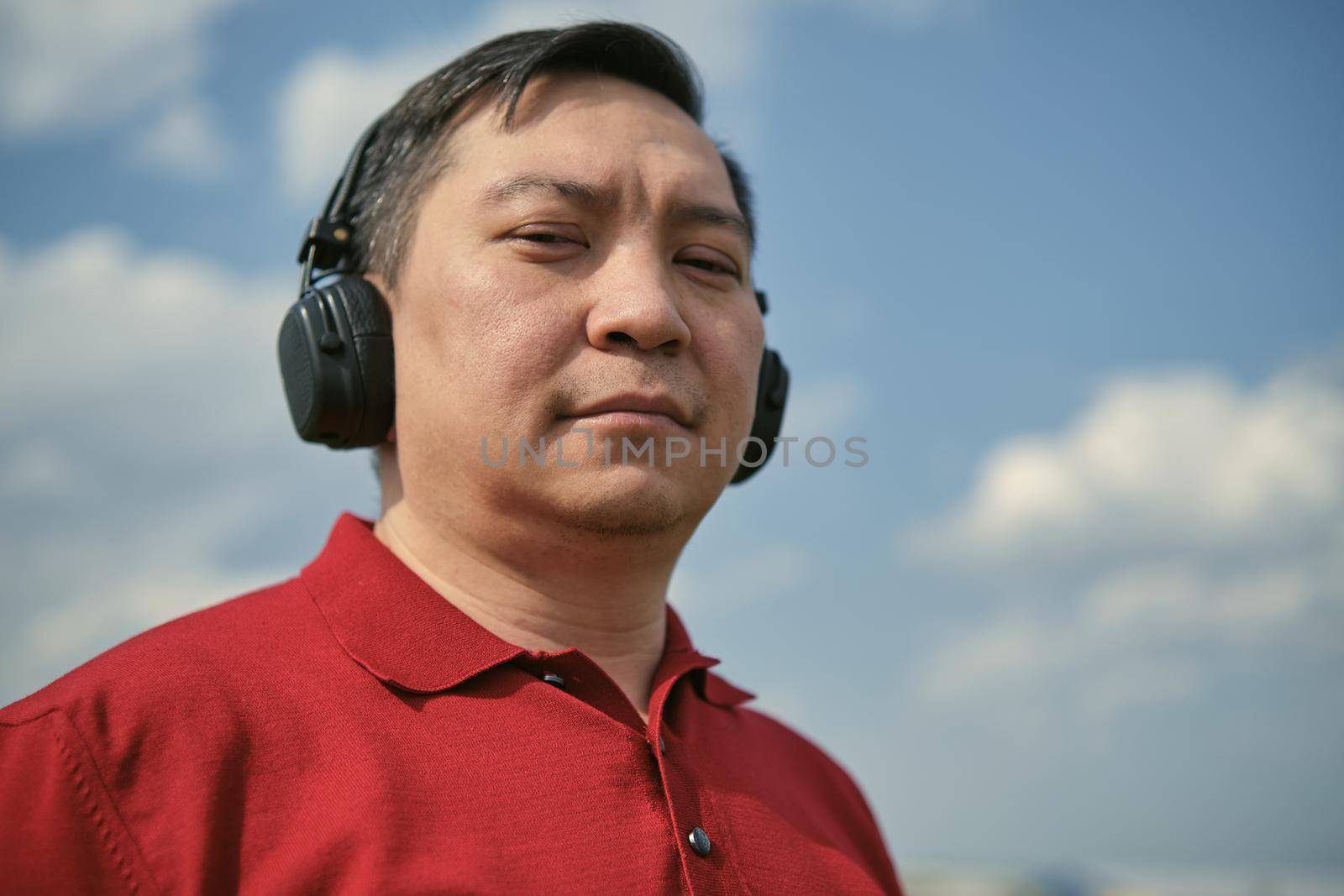 Middle-aged Asian man in headphones outdoors listening to music against the background of the sky