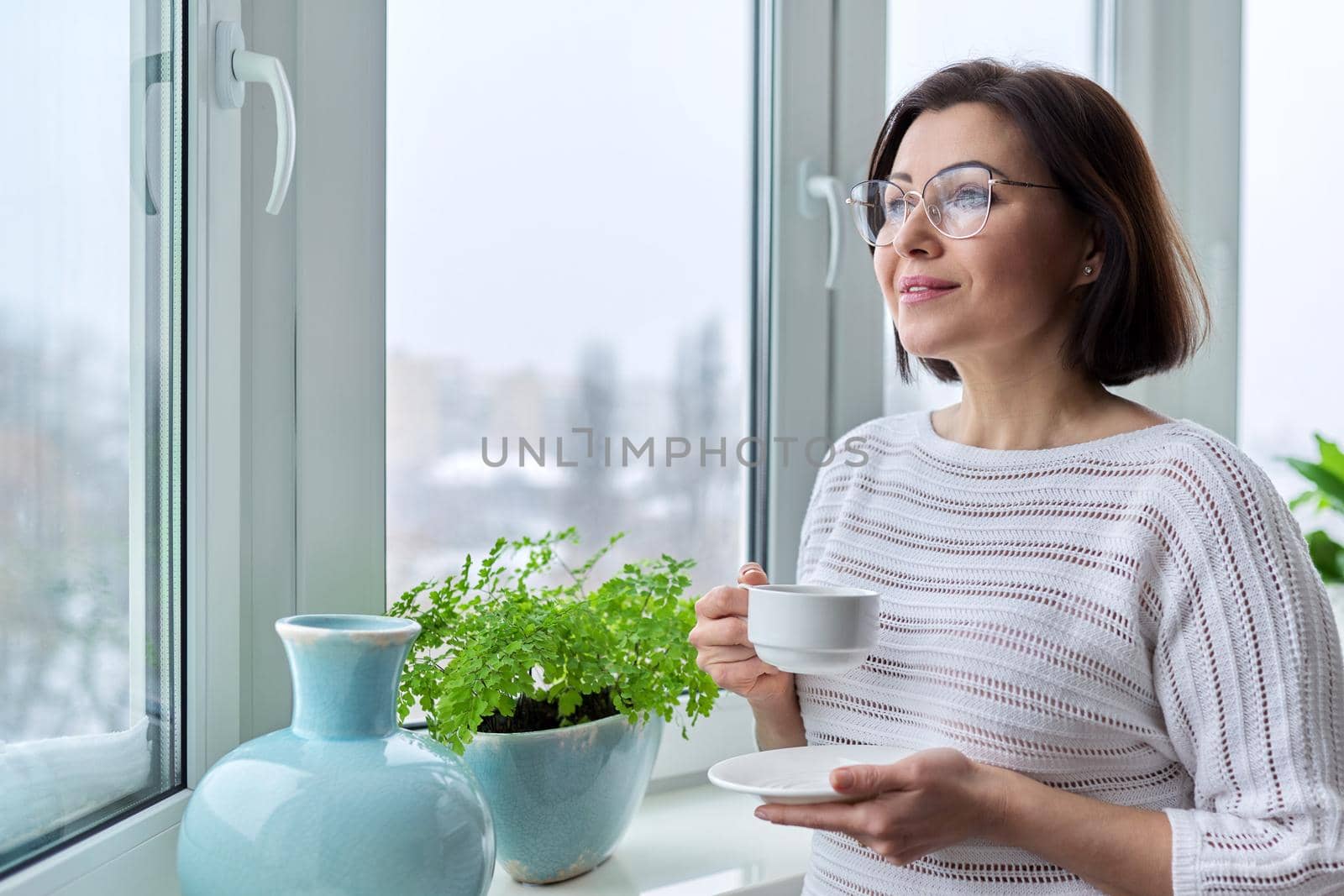 Middle aged beautiful woman with glasses with a cup of coffee at home near the window in the winter autumn season, copy space. Beauty, lifestyle, comfort, tranquility, mature age concept