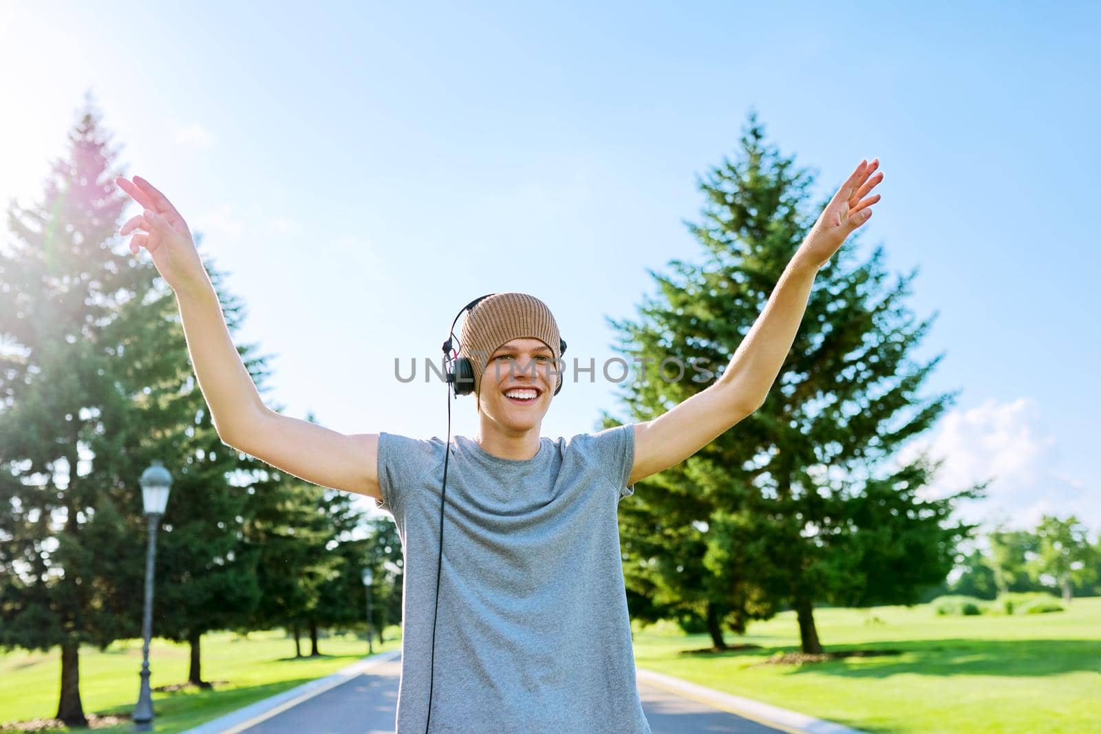Portrait of cheerful hipster guy in knitted hat with headphones, dancing, hands up. Smiling teenager listening to music, summer in park. Hobbies, leisure, creativity, youth, lifestyle and technology