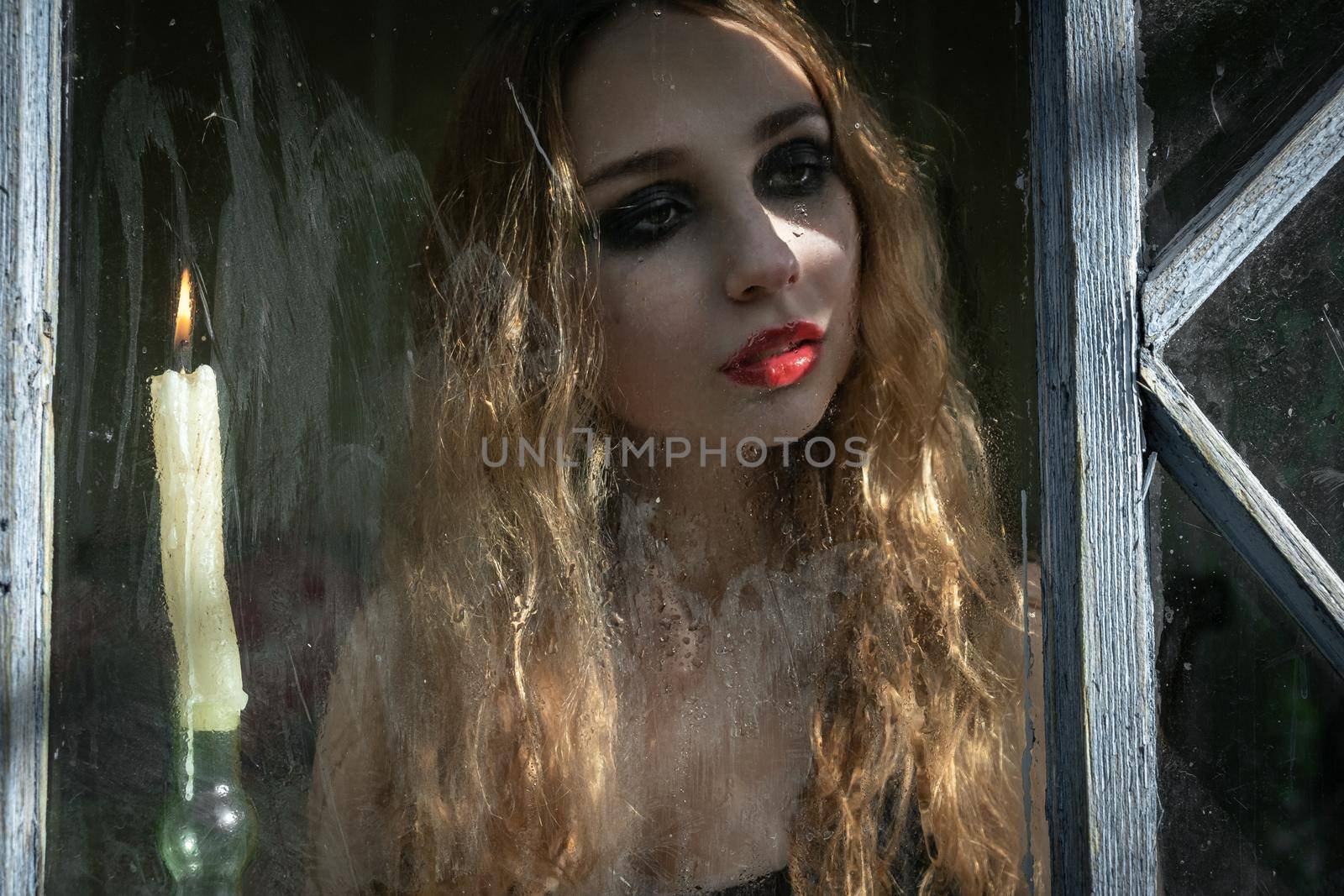 Beautiful sad young girl looking out the window with a candle, good for book cover