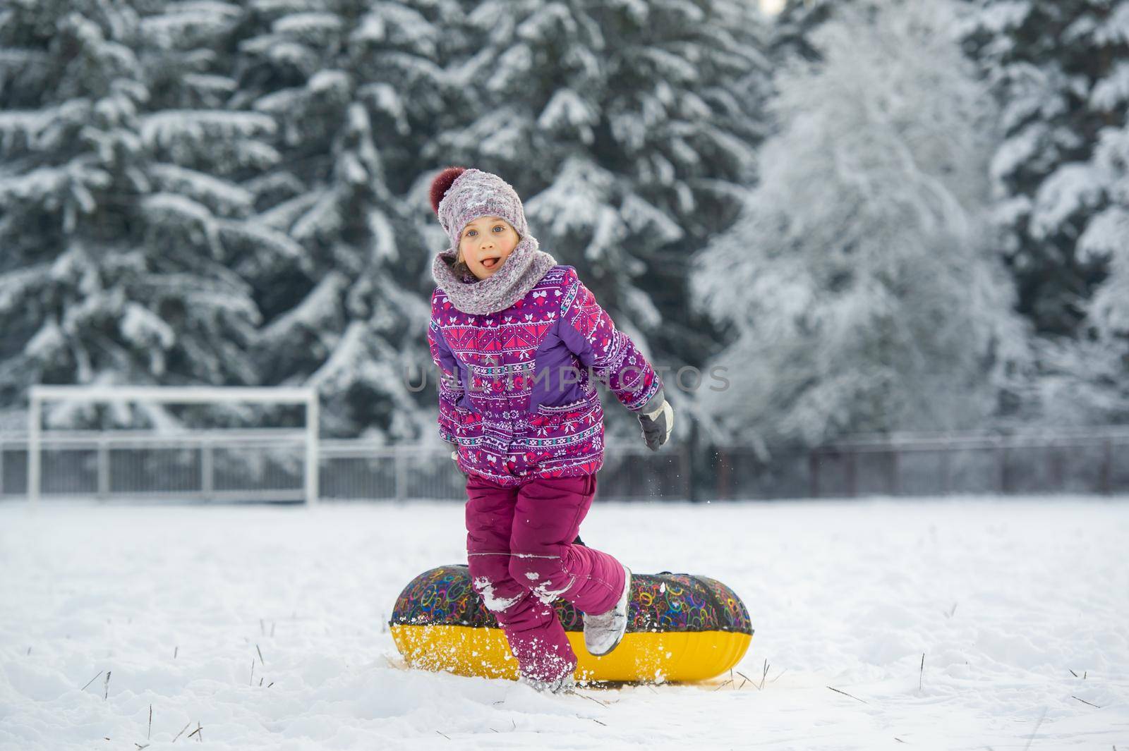 a little girl in winter in purple clothes and an inflatable circle walks on the street in a snow-covered forest.