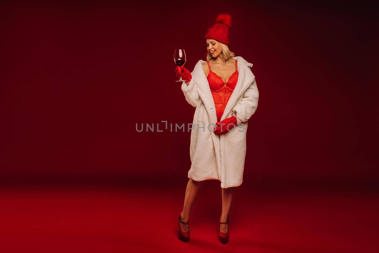 portrait of a smiling girl in a white fur coat and underwear holding a glass of champagne on a red background.