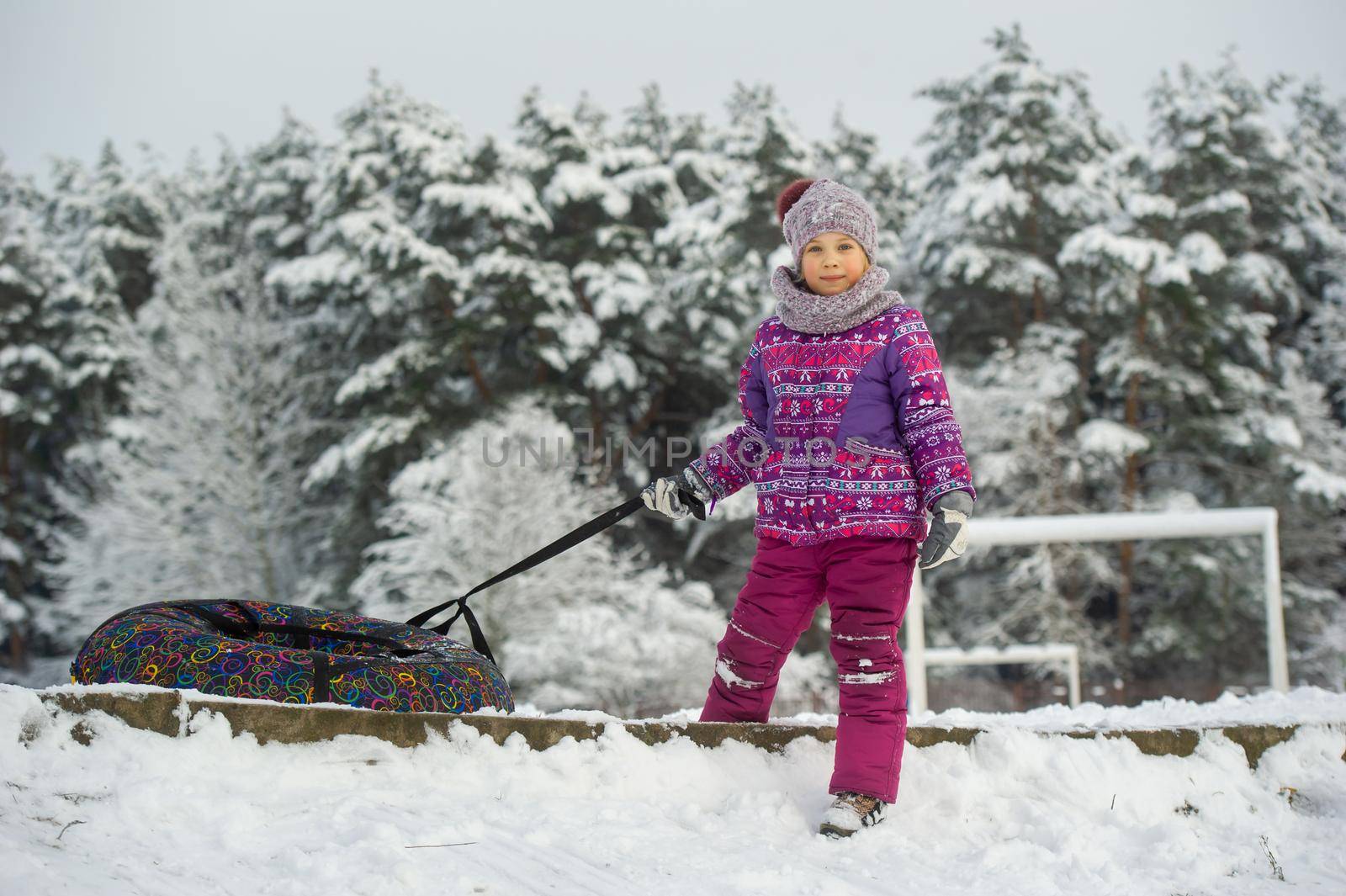 a little girl in winter in purple clothes and an inflatable circle walks on the street in a snow-covered forest.