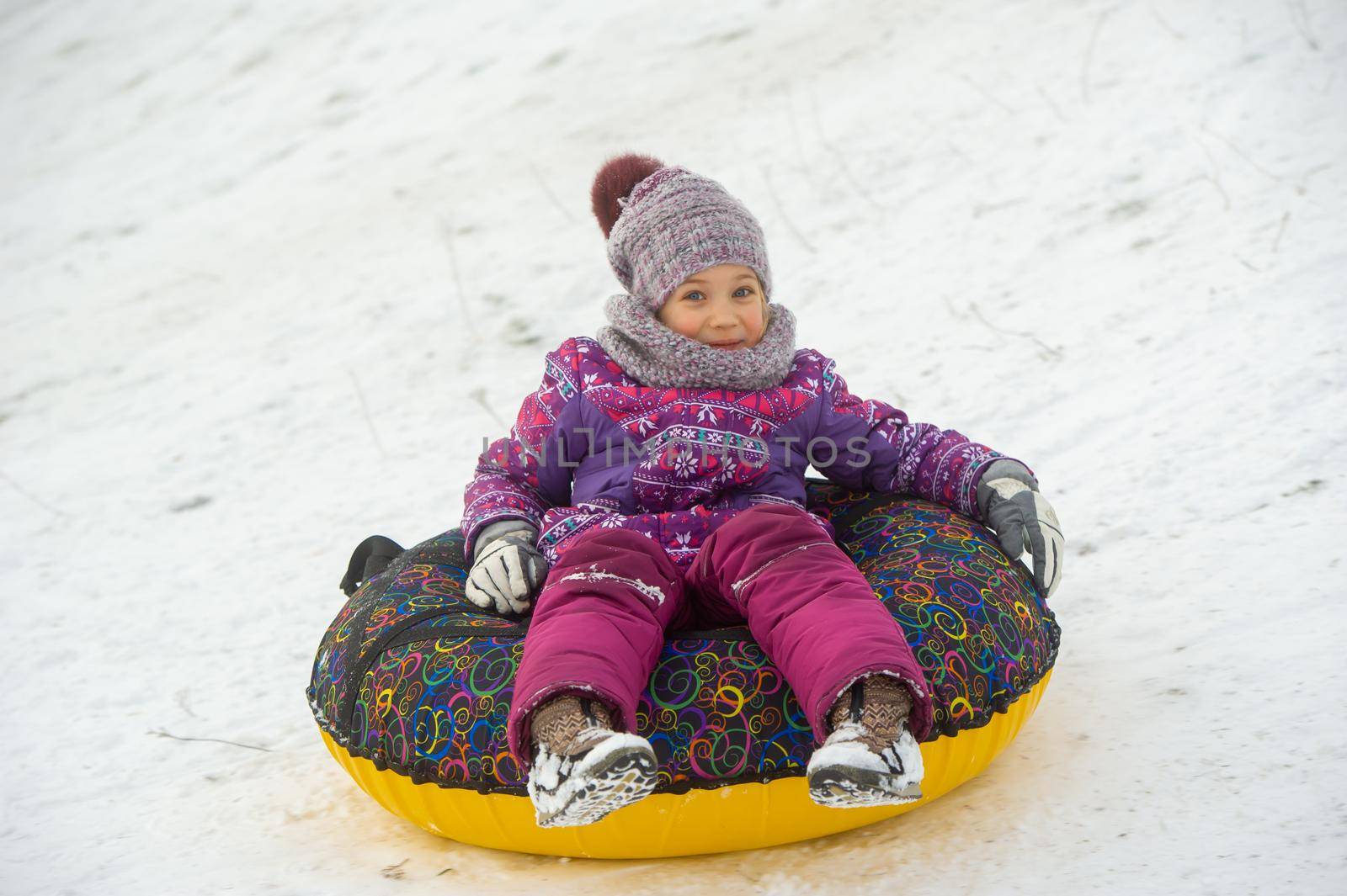 a little girl in winter in purple clothes and an inflatable circle rides down the hill on the street.