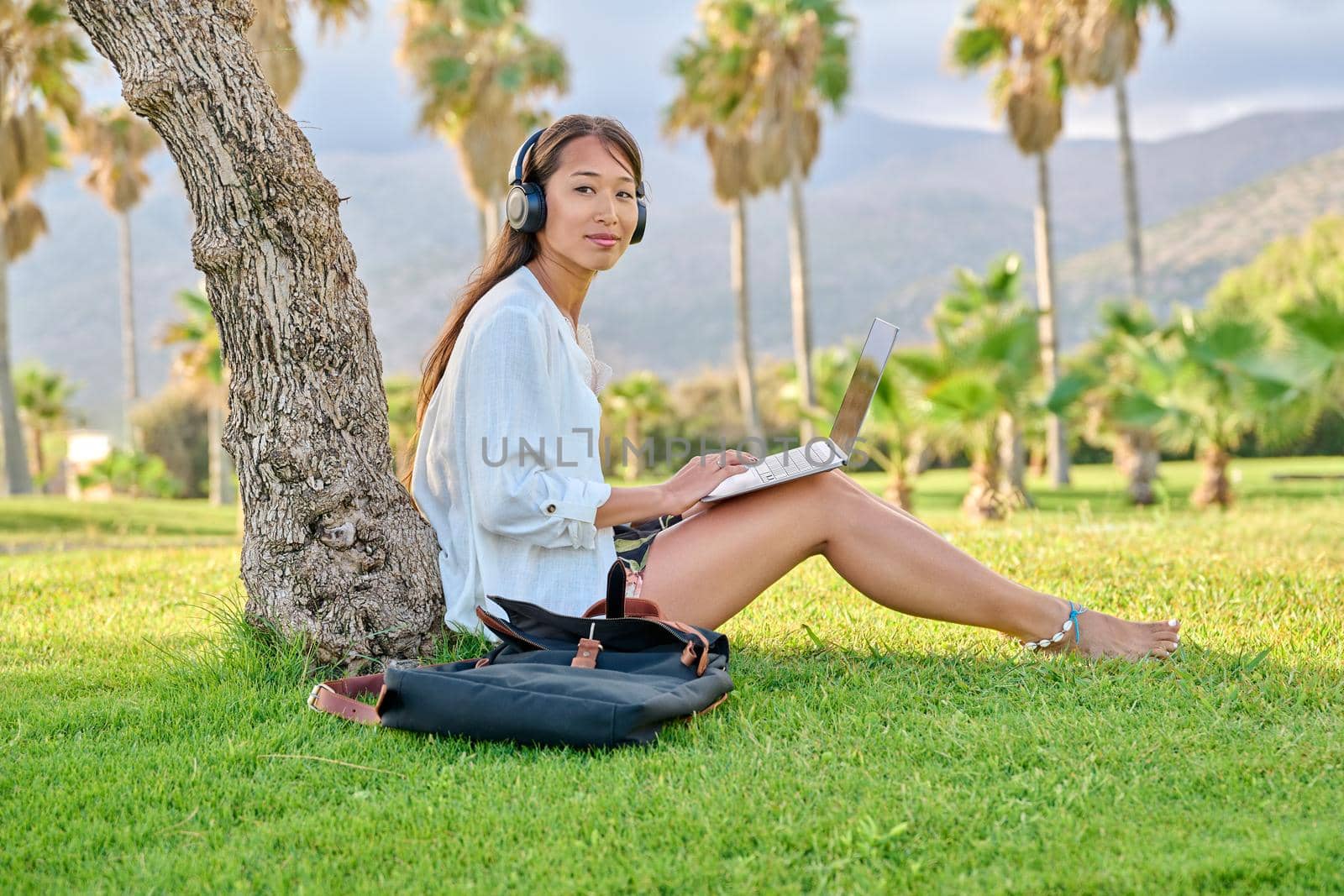 Young woman in headphones working on laptop outdoors. Asian female university student studying remotely, creative freelancer, business woman, blogger vlogger, work and study online, modern technology