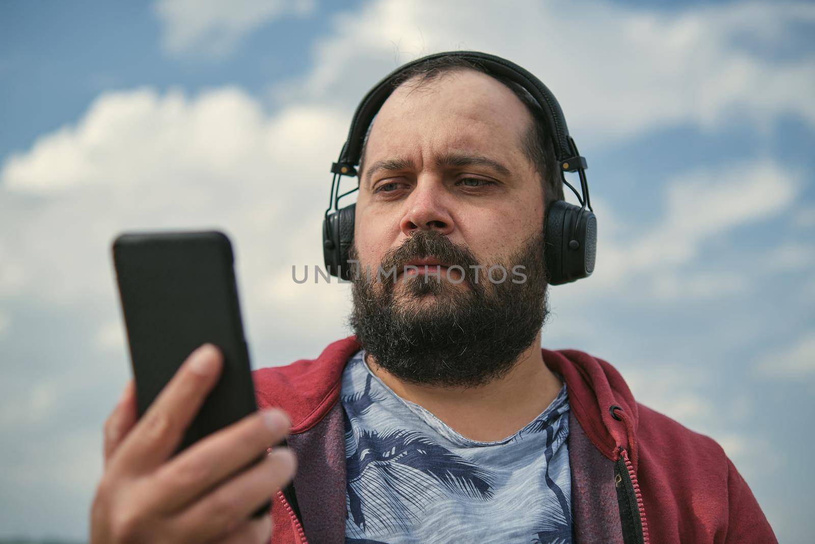 Middle-aged European man in headphones outdoors listening to music against the background of the sky, mobile phone in his hand
