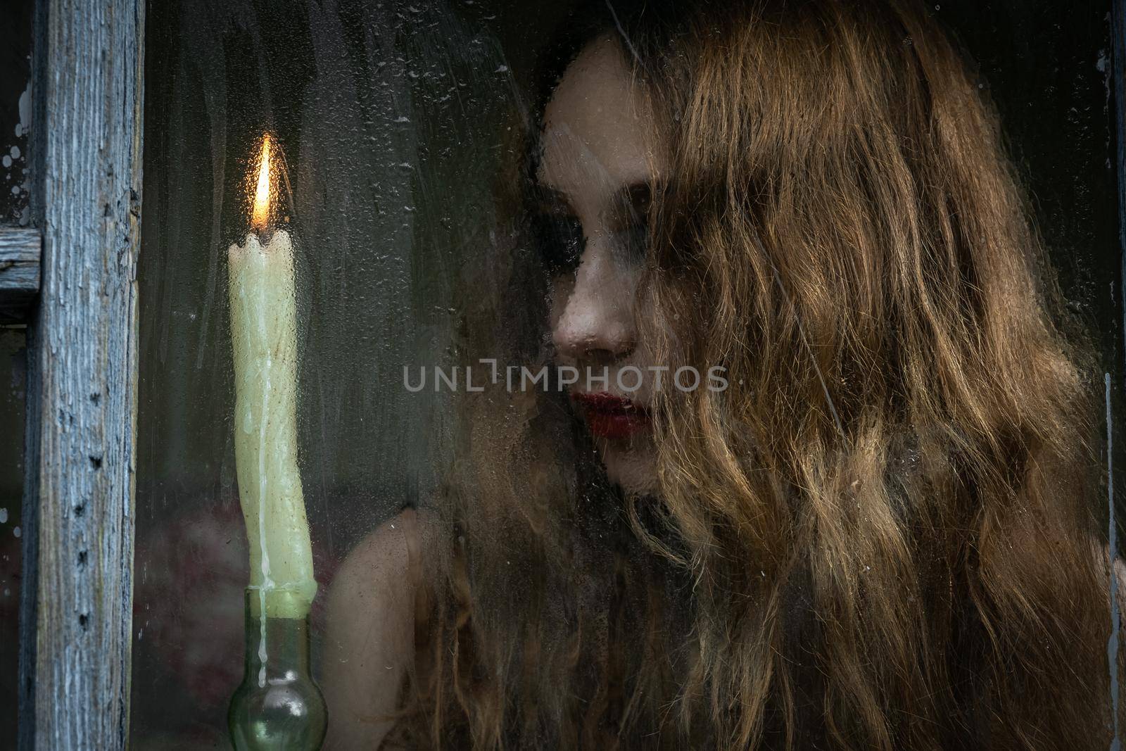 Beautiful sad young girl looking out the window with a candle, good for book cover