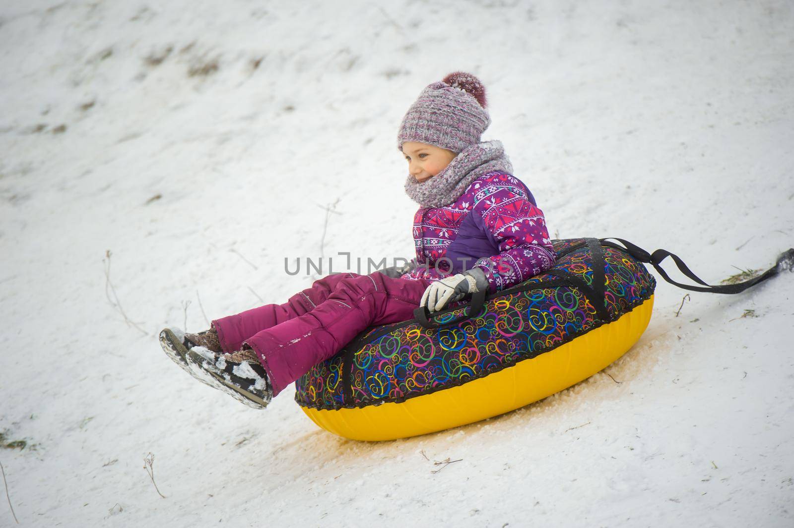 a little girl in winter in purple clothes and an inflatable circle rides down the hill on the street.