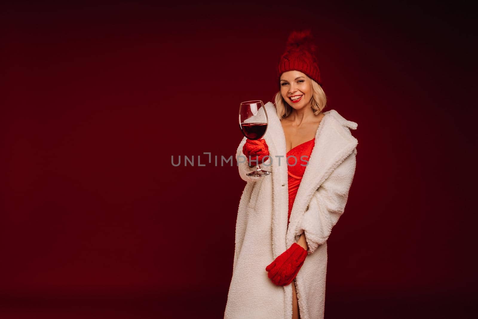 portrait of a smiling girl in a white fur coat and underwear holding a glass of champagne on a red background.