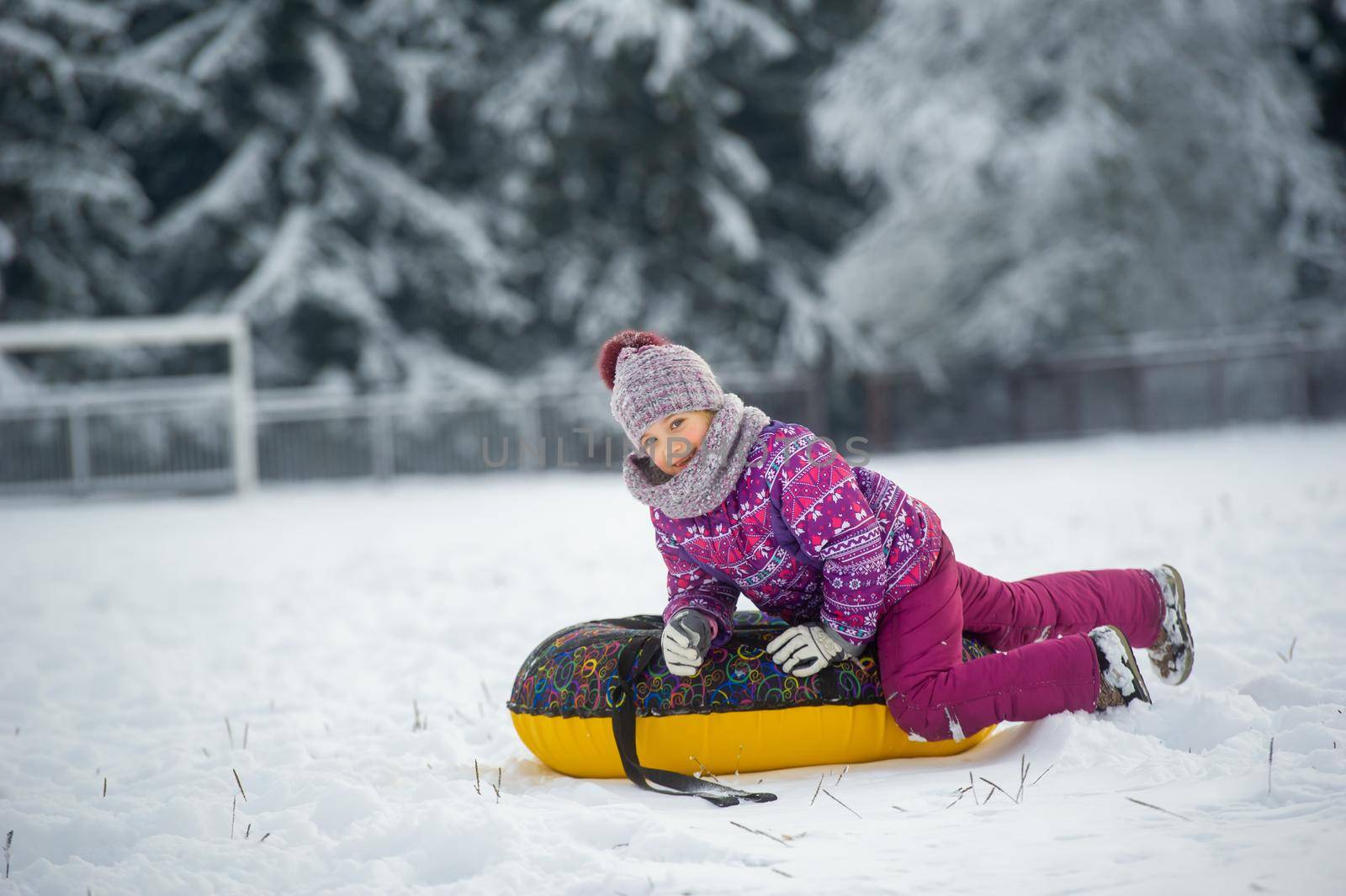 a little girl in winter in purple clothes and an inflatable circle walks on the street in a snow-covered forest.