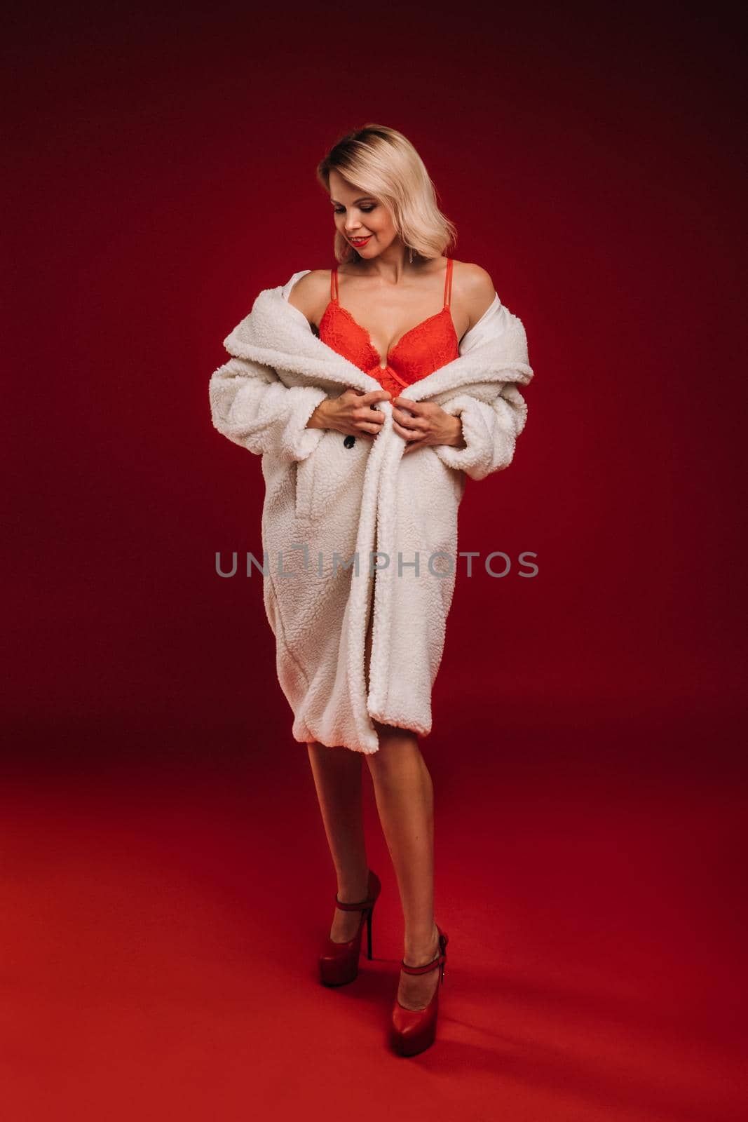 portrait of a smiling girl in a white fur coat and underwear holding a glass of champagne on a red background.