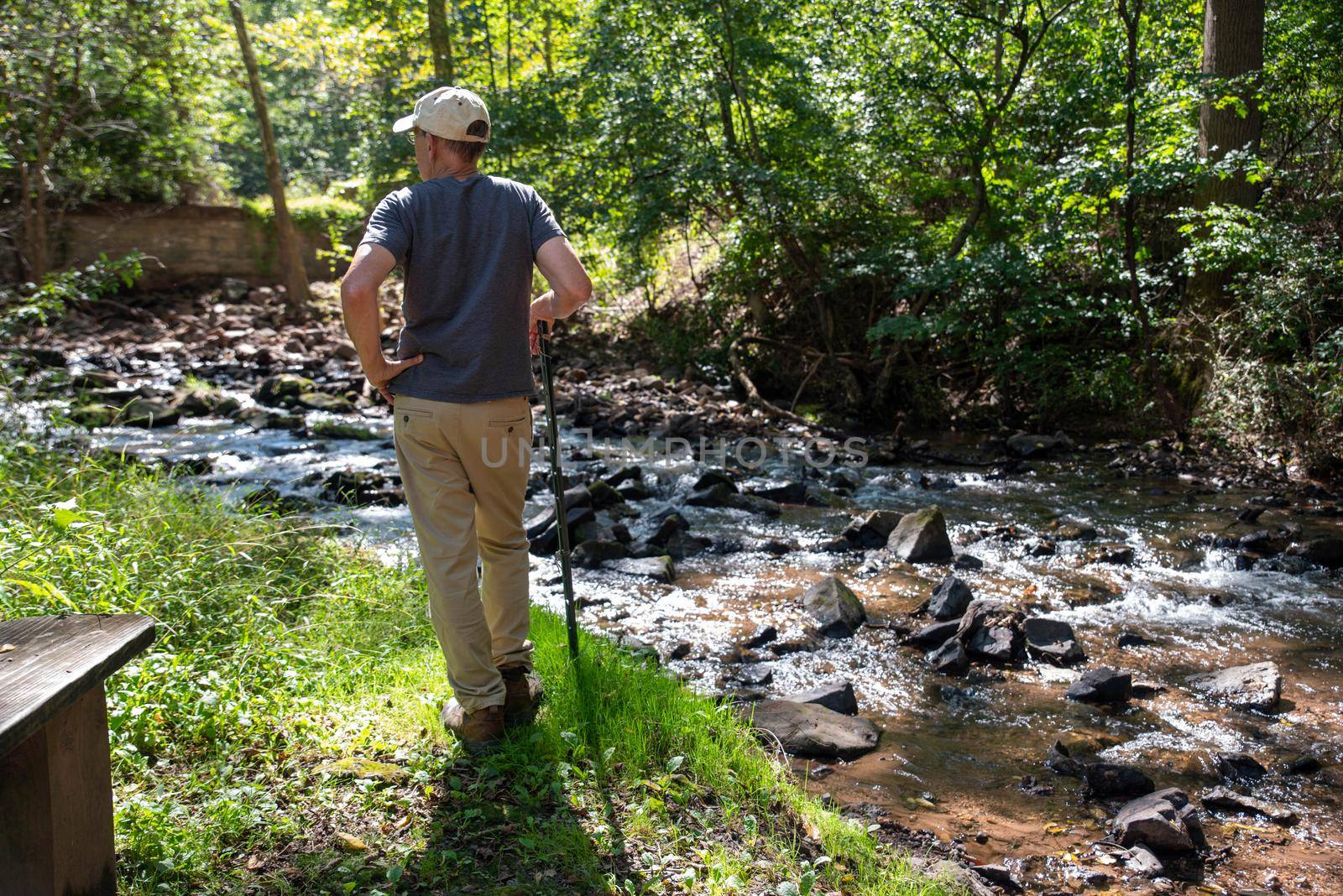 Active senior man stops and contemplates nature in meditative setting with flowing stream and green woodland background. Serene image shot in natural light with one man and copy space