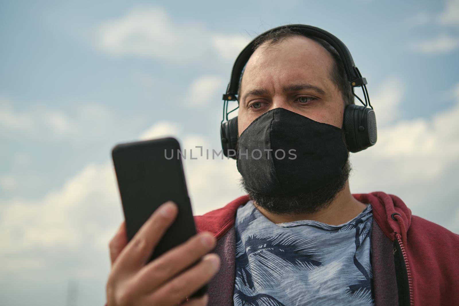 Middle-aged European man in headphones outdoors listening to music against the background of the sky, mobile phone in his hand