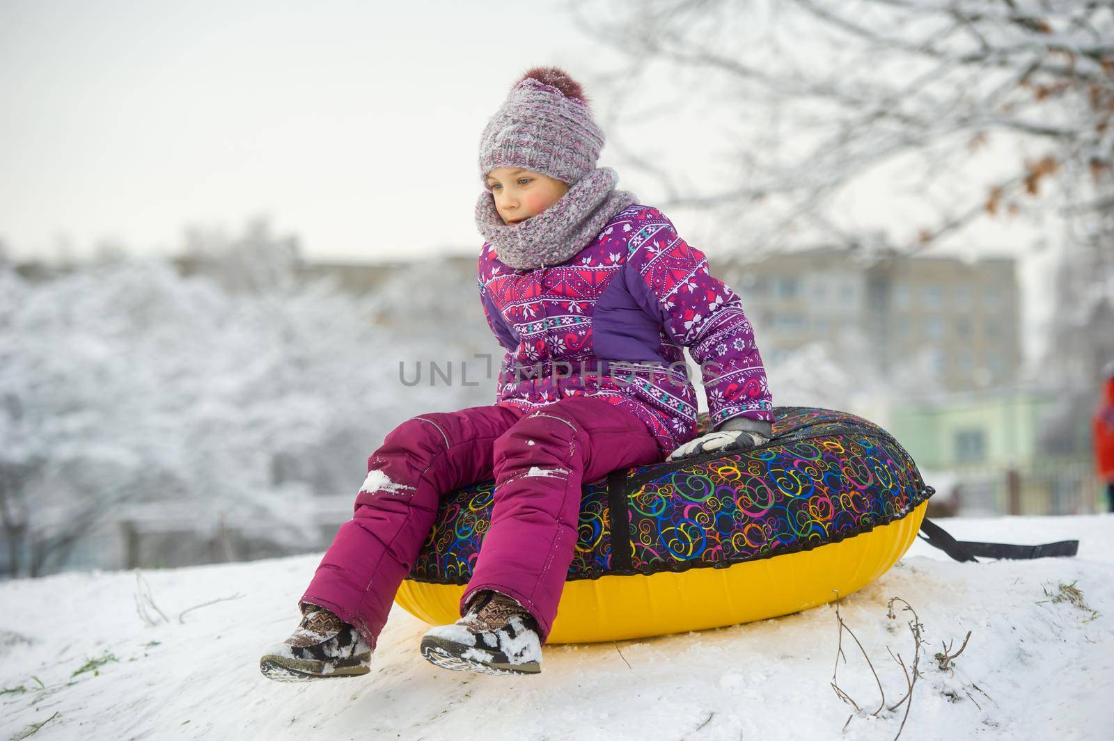 a little girl in winter in purple clothes and an inflatable circle rides down the hill on the street.