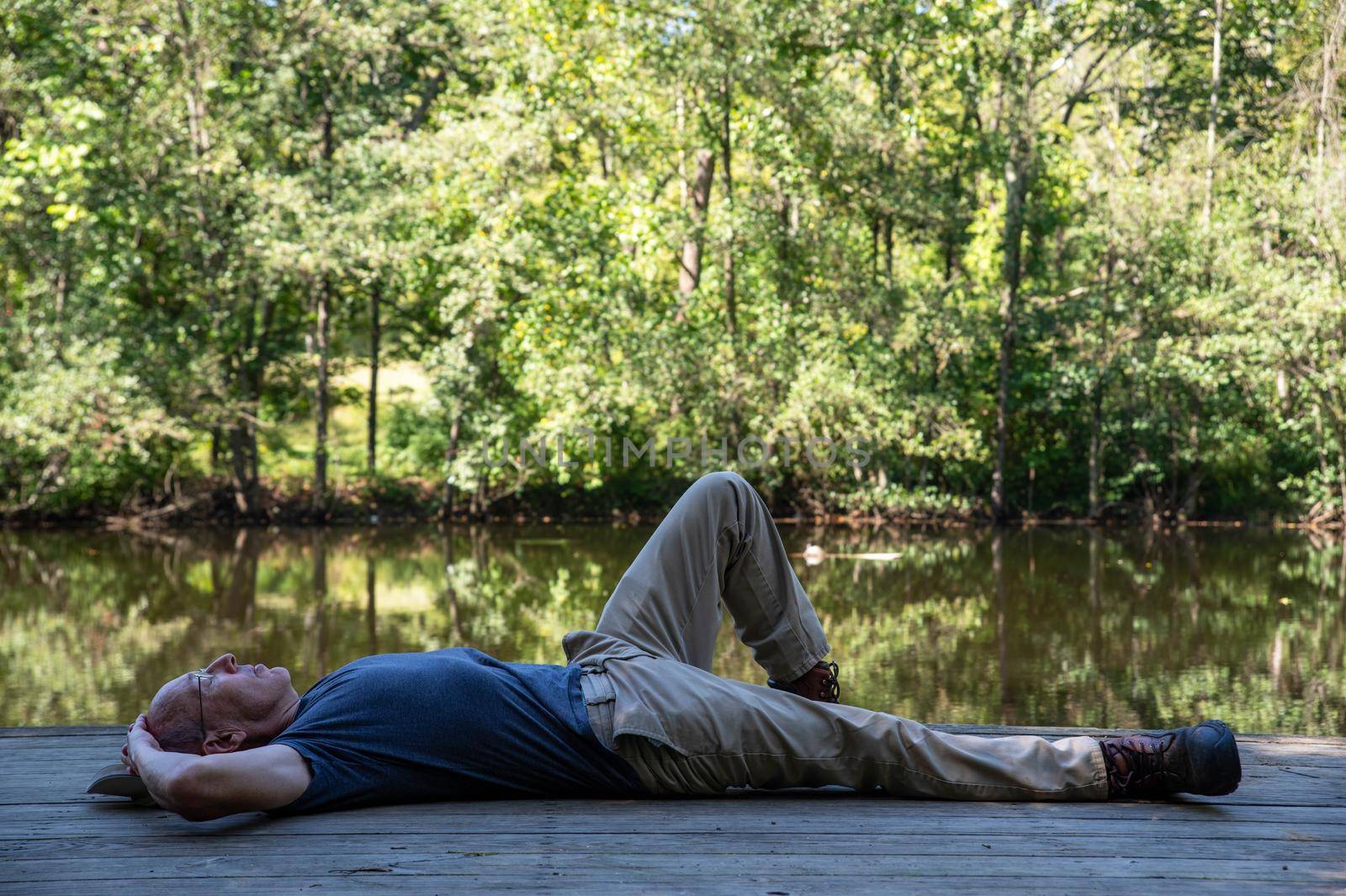 Caucasian senior man lying on a wooden dock alone with his eyes closed by a serene woodland pond with green bokeh nature background