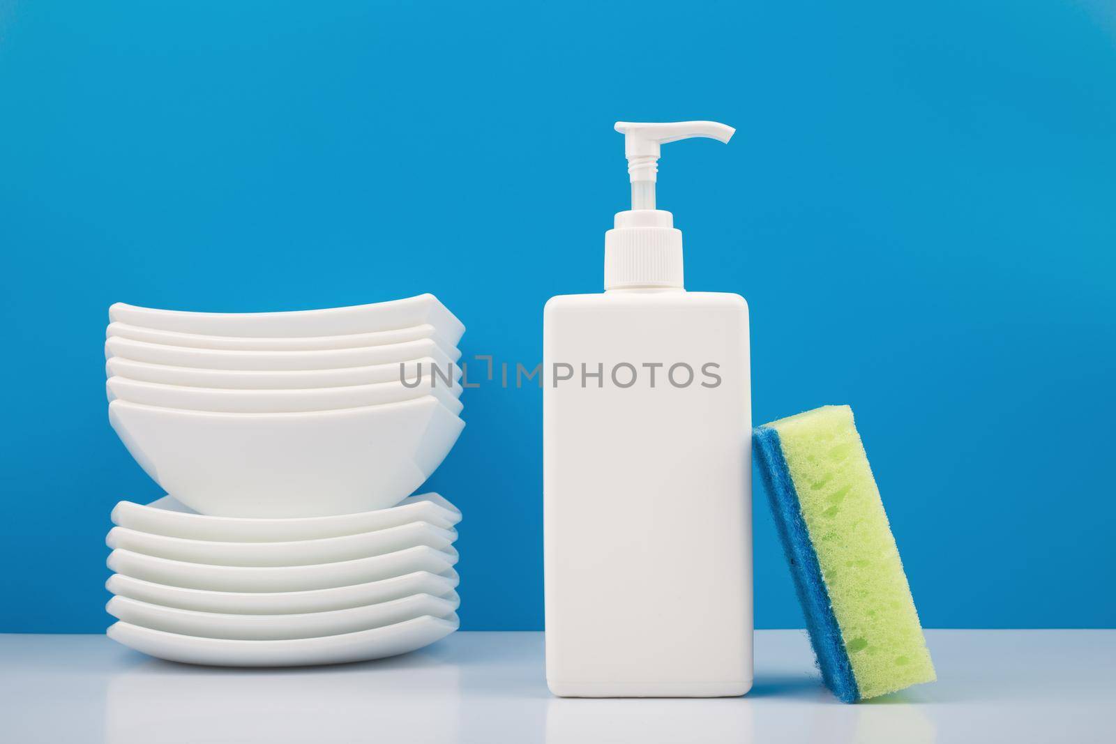 Colorful dishwashing concept, creative composition with liquid detergent in white plastic bottle with dispenser, green cleaning sponge and pile of clean plates on white table against blue background