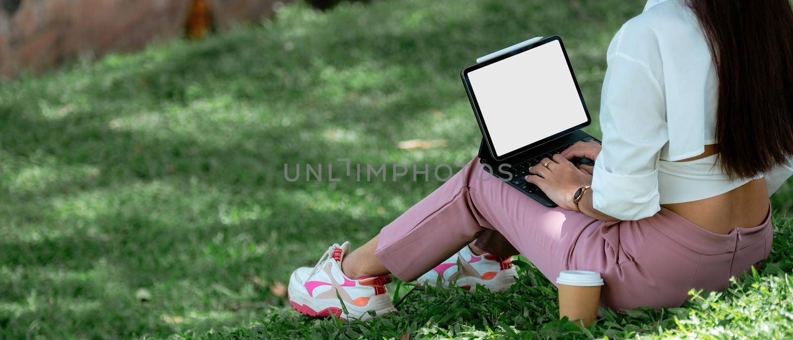 Close up student with digital tablet blank screen sitting on green grass lawn writing in notebook, technology in education, college, high school, teens concept. by nateemee