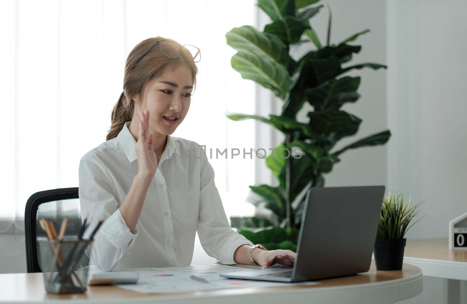 Smiling young female employee at home hand wave on video call on laptop with diverse colleagues. Asian woman worker have webcam conference or digital web team meeting or briefing with coworkers.