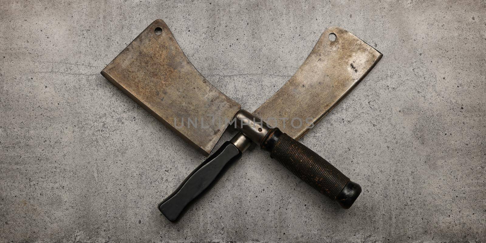 Close up two crossed vintage butcher meat cleavers on cutting board or grunge gray stone table surface with copy space, elevated top view, directly above